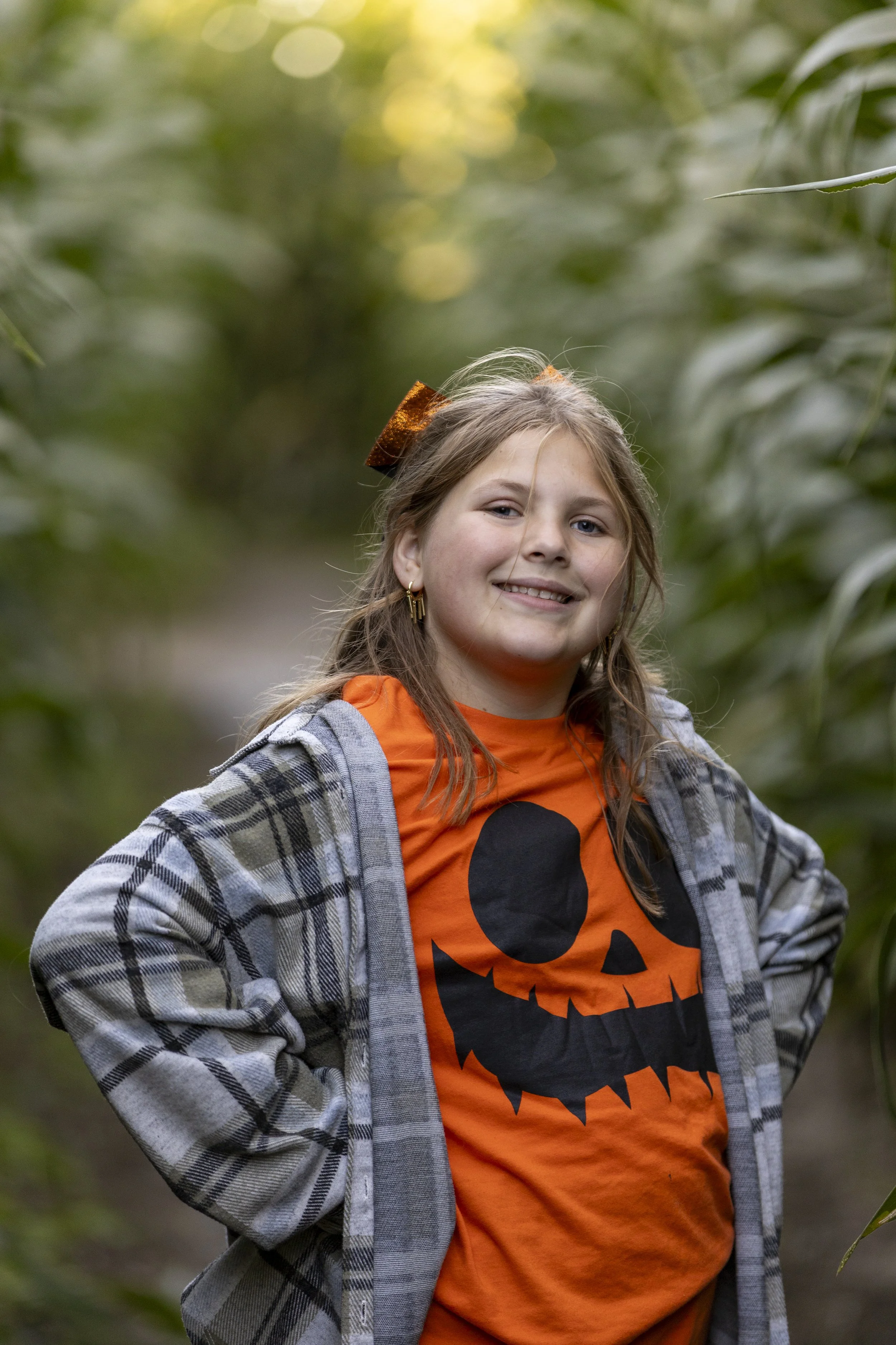 A young girl smiling at the camera, wearing an orange Halloween pumpkin shirt, a plaid jacket, and a hair bow, standing outdoors among green plants.