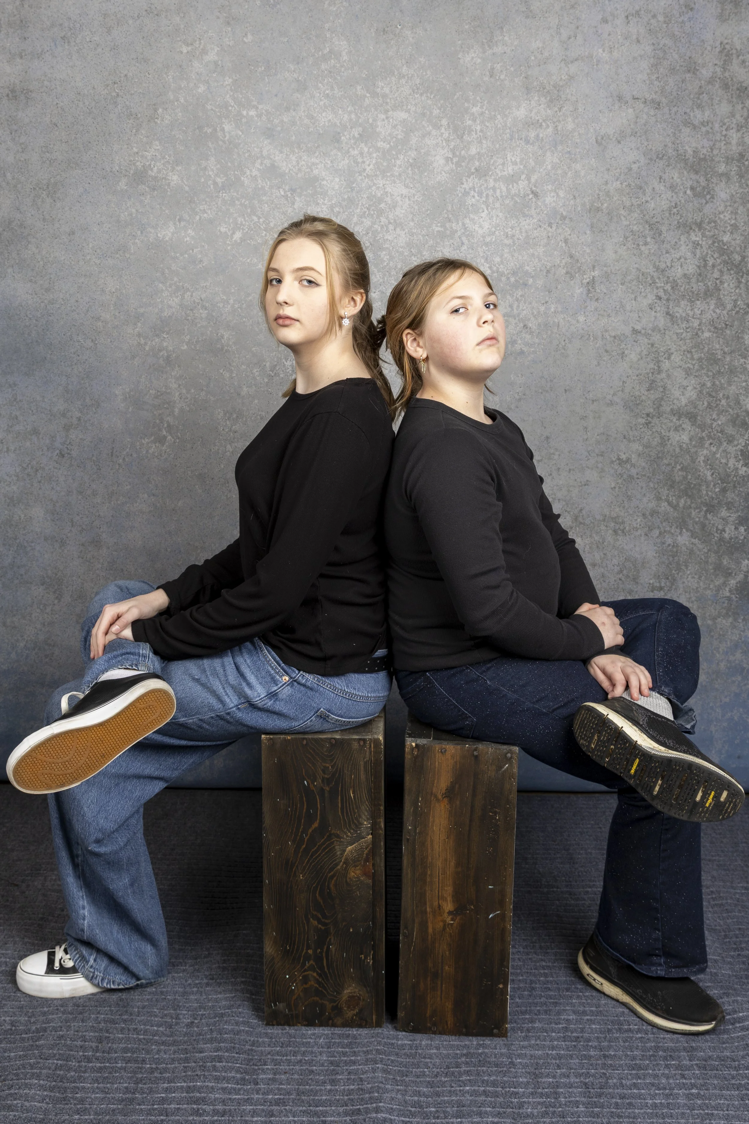 Two girls sitting back-to-back on wooden blocks, with neutral expressions, against a gray textured background. Both are wearing black long-sleeve shirts and jeans.