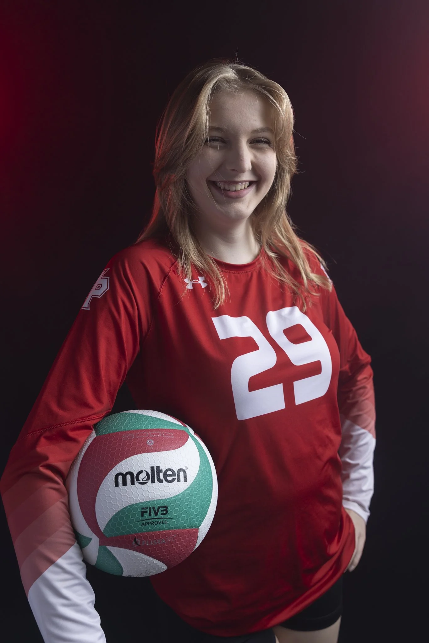 Young woman with light brown hair wearing a red volleyball jersey with the number 29, holding a volleyball, smiling, dark background with pinkish-purple lighting.