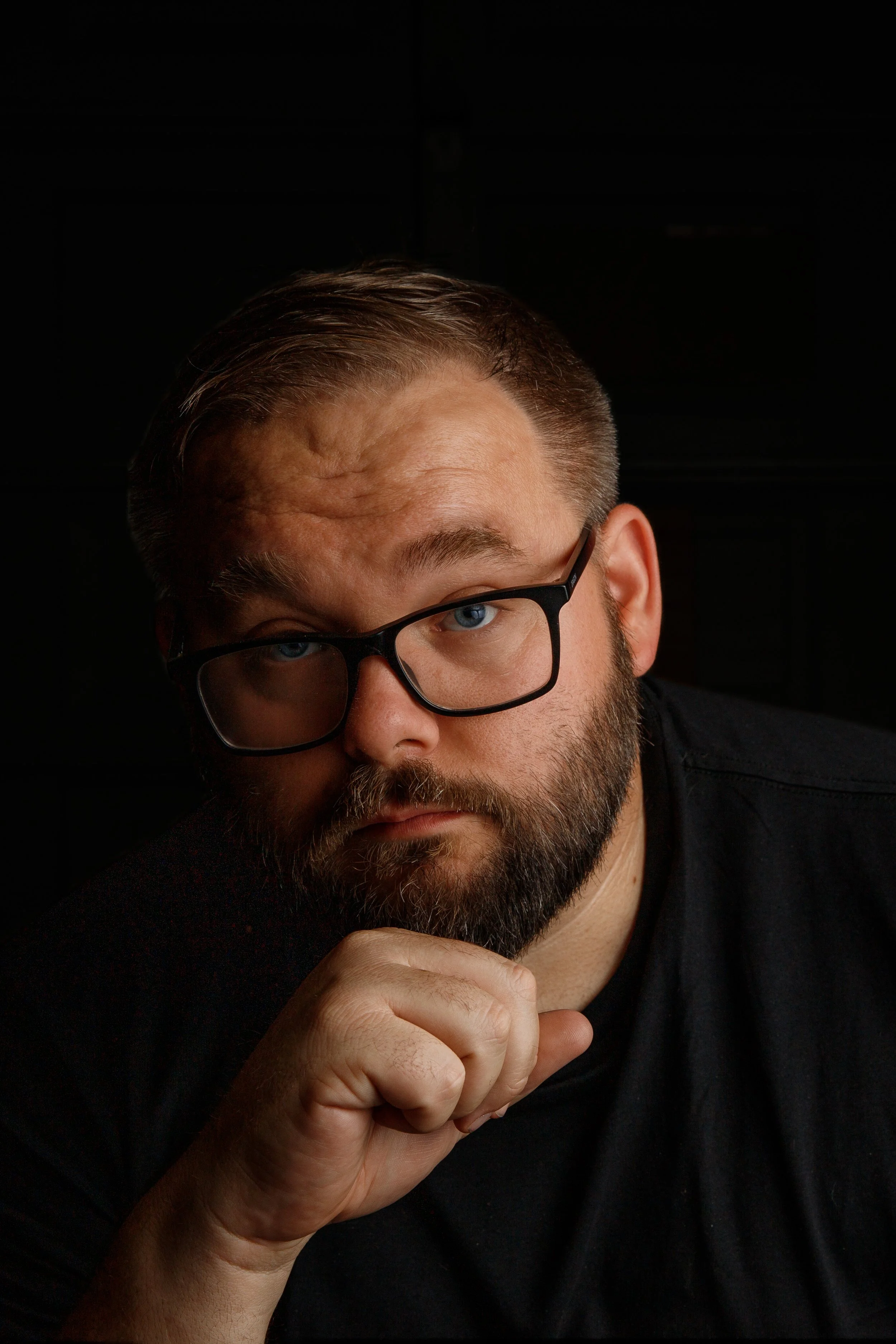 A man with glasses and a beard looking thoughtfully at the camera, with his hand resting near his chin, against a dark background.