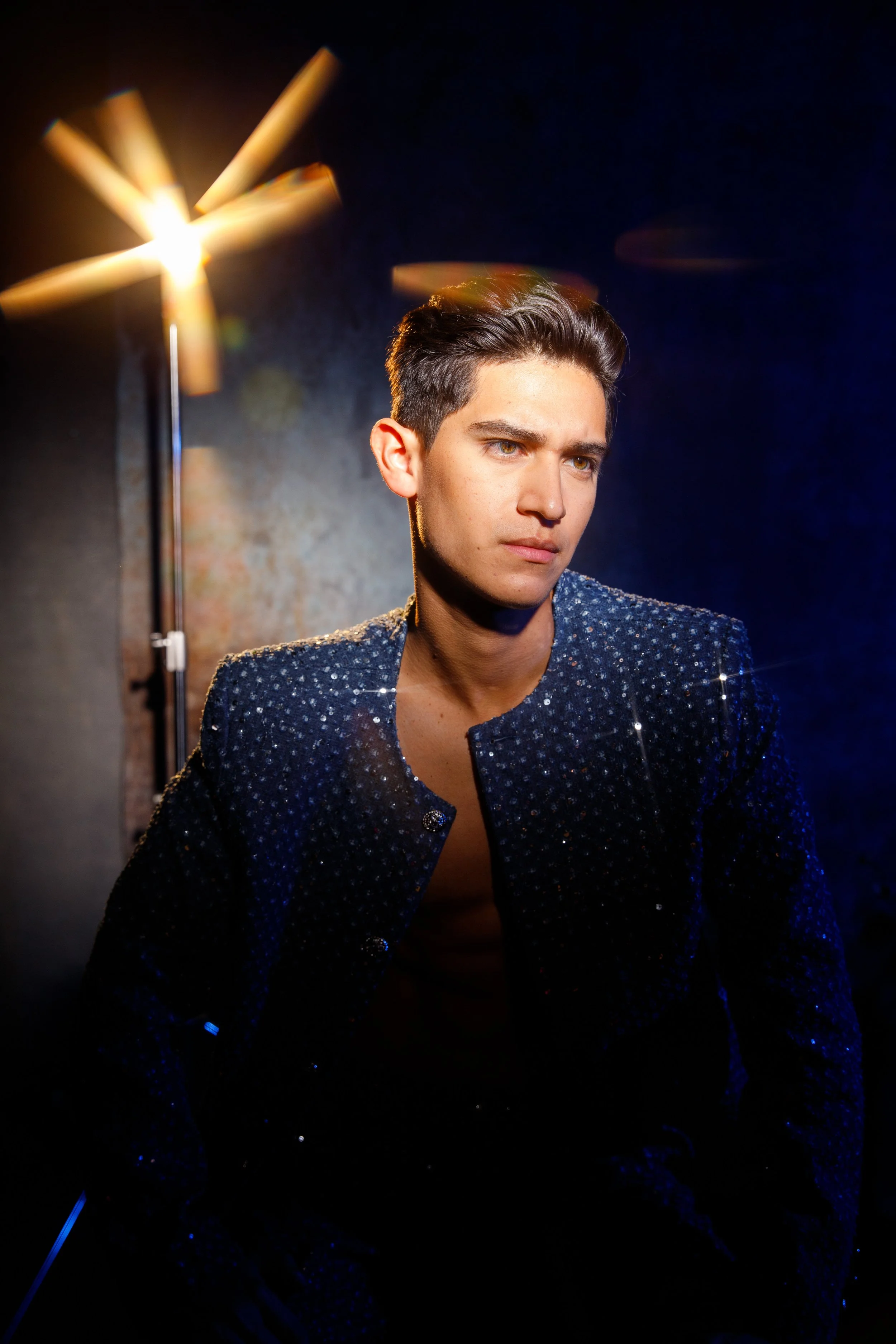 A young man with dark, styled hair and light skin posing in a dark setting, wearing a sparkly, dark blue jacket and standing in front of a light source creating a glow effect.