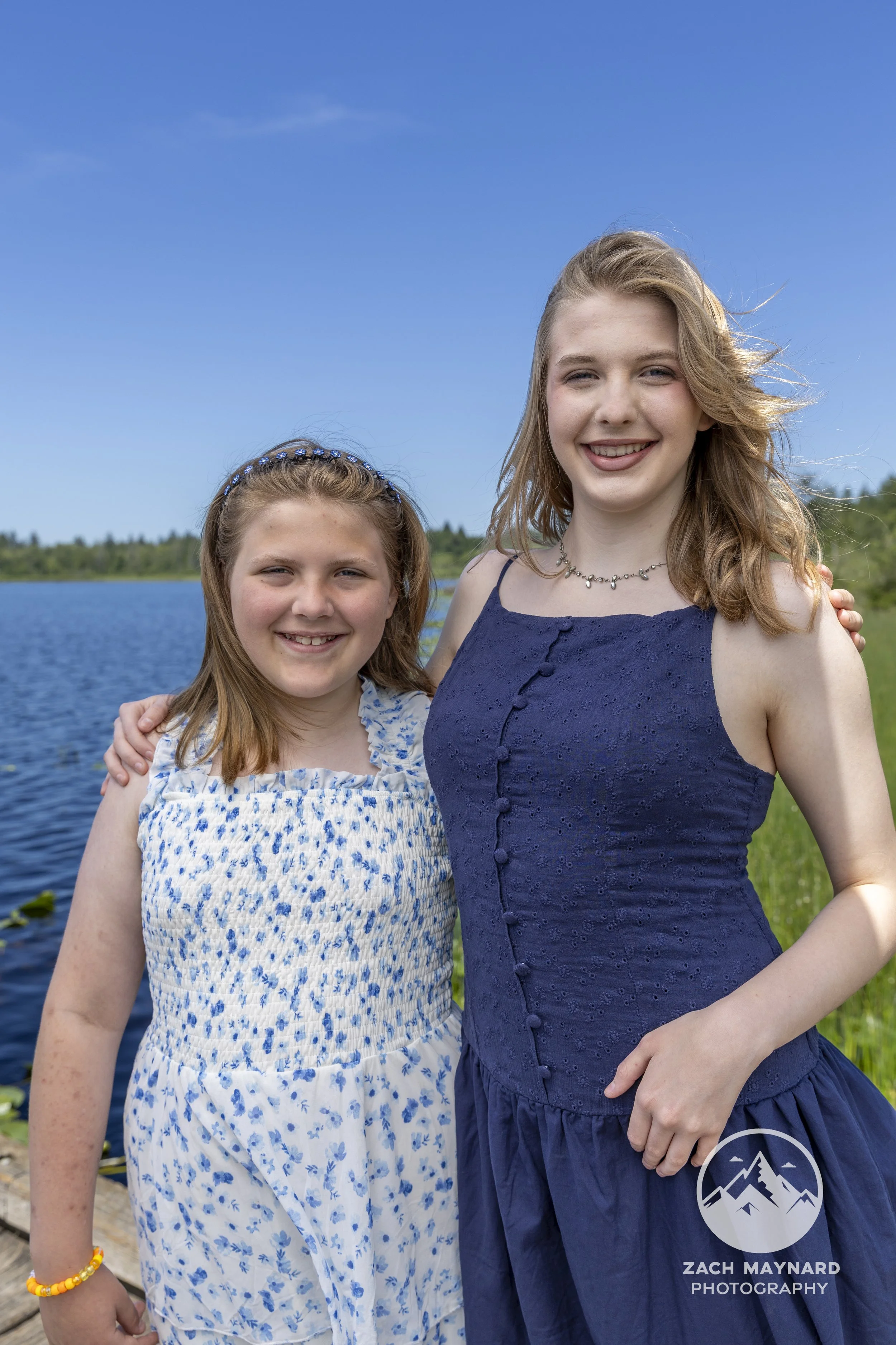 Two sisters in floral and navy dresses smiling lakeside during a summer family session in Snohomish County