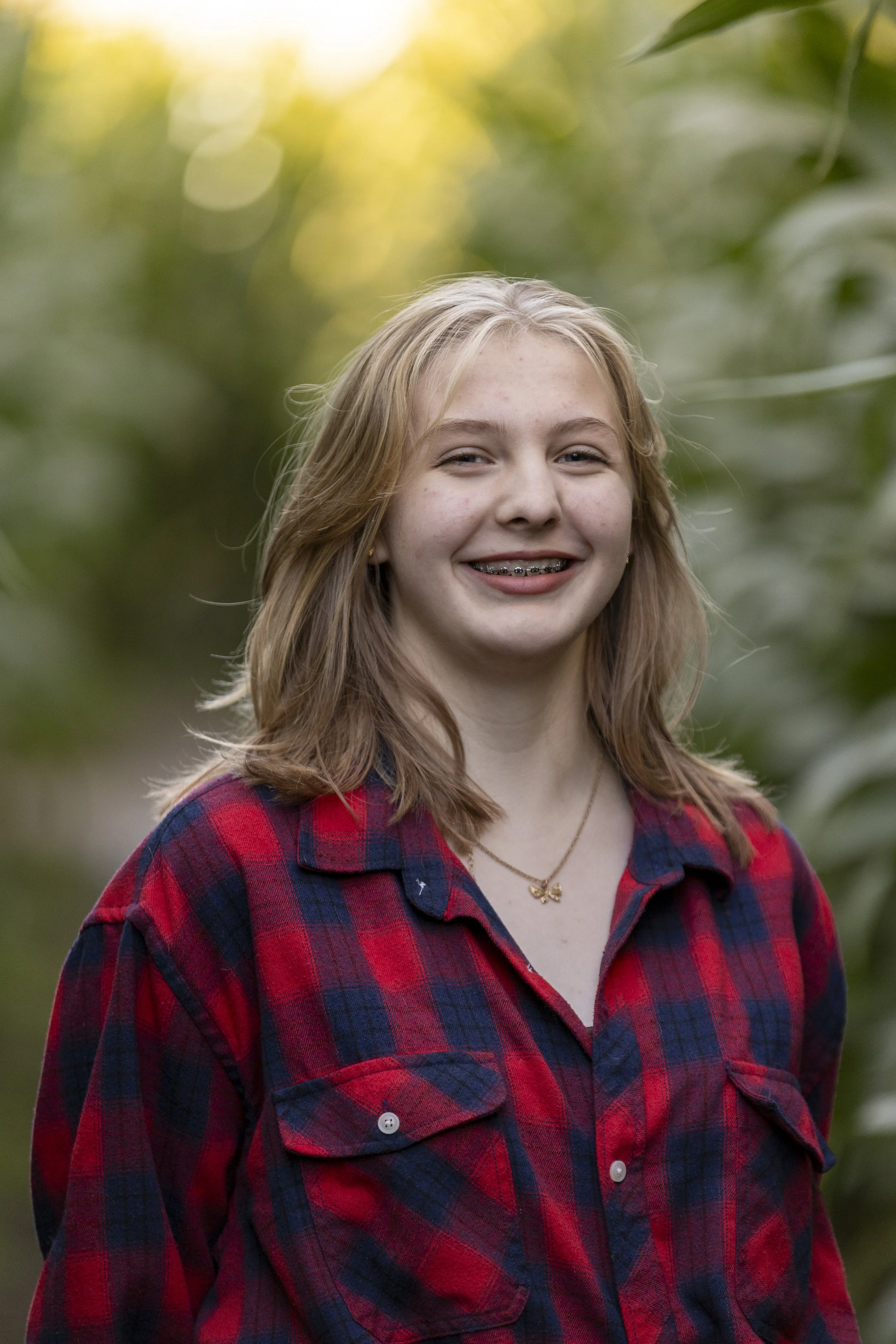 A young girl with light brown hair, braces, and a red and blue plaid shirt, smiling outdoors with blurred green foliage in the background.