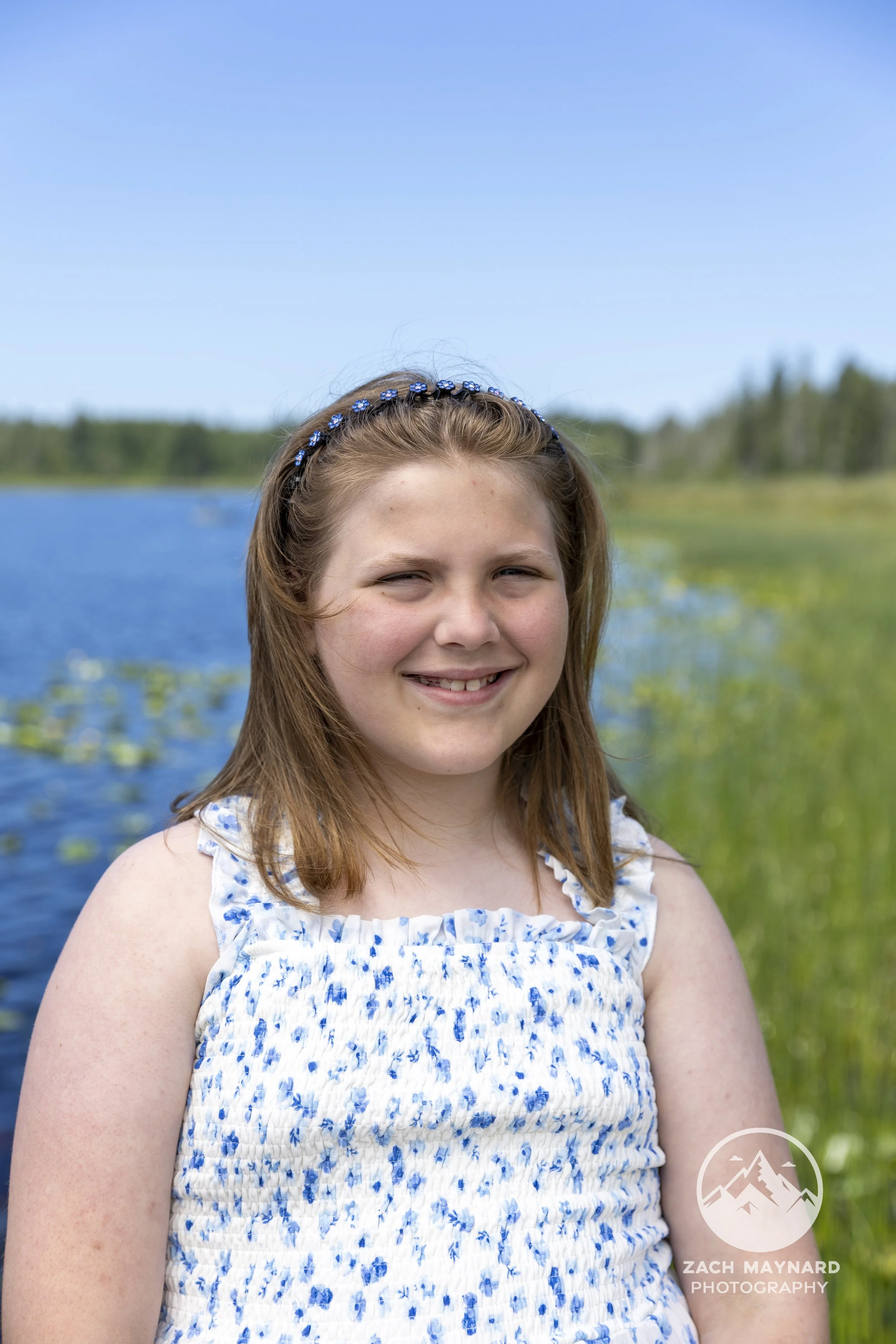 Young girl in a blue floral dress smiling by a lake during a summer portrait session in Snohomish County