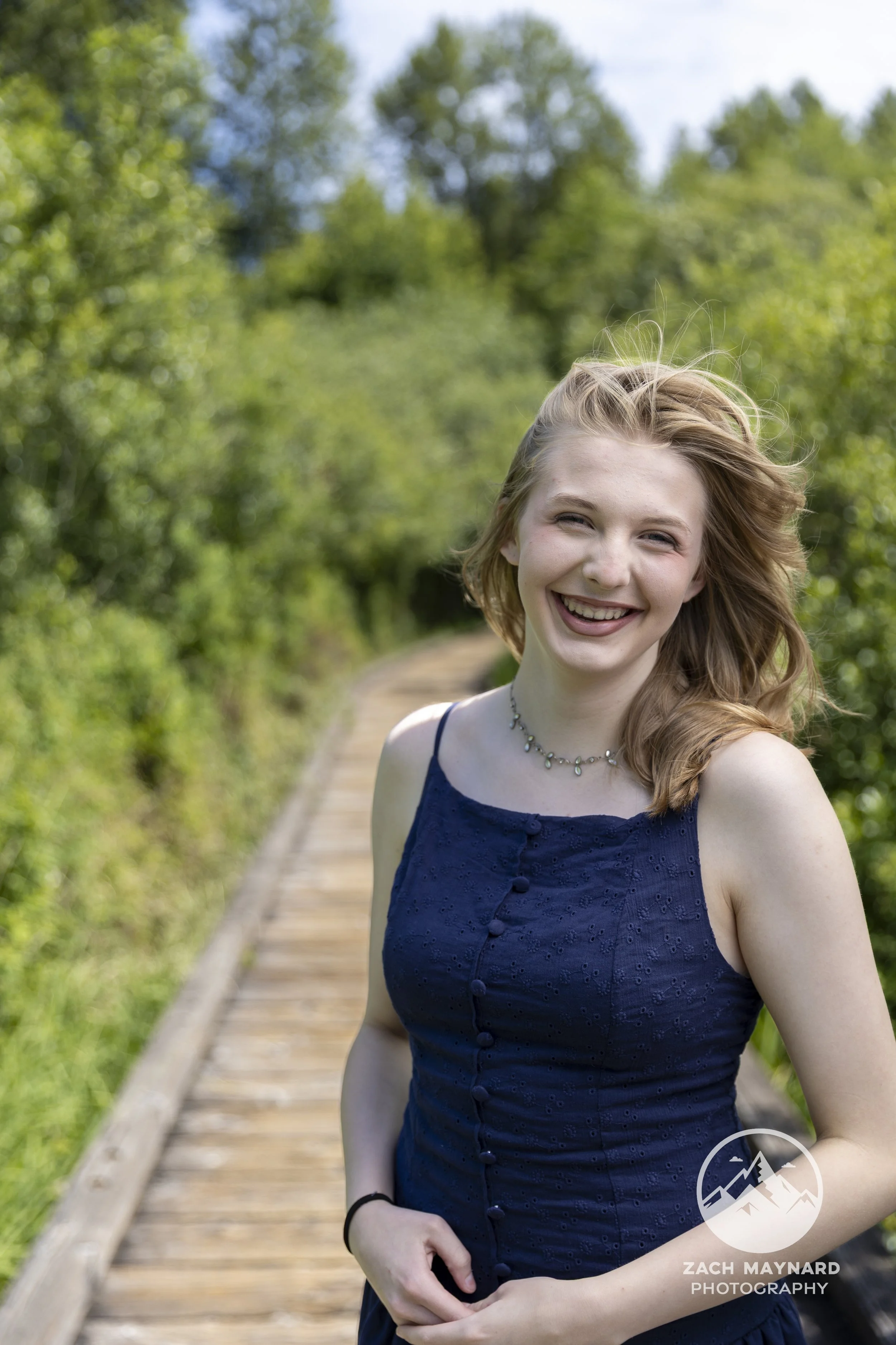 Teen girl laughing in a navy dress on a forest boardwalk during a summer senior portrait session in Snohomish County