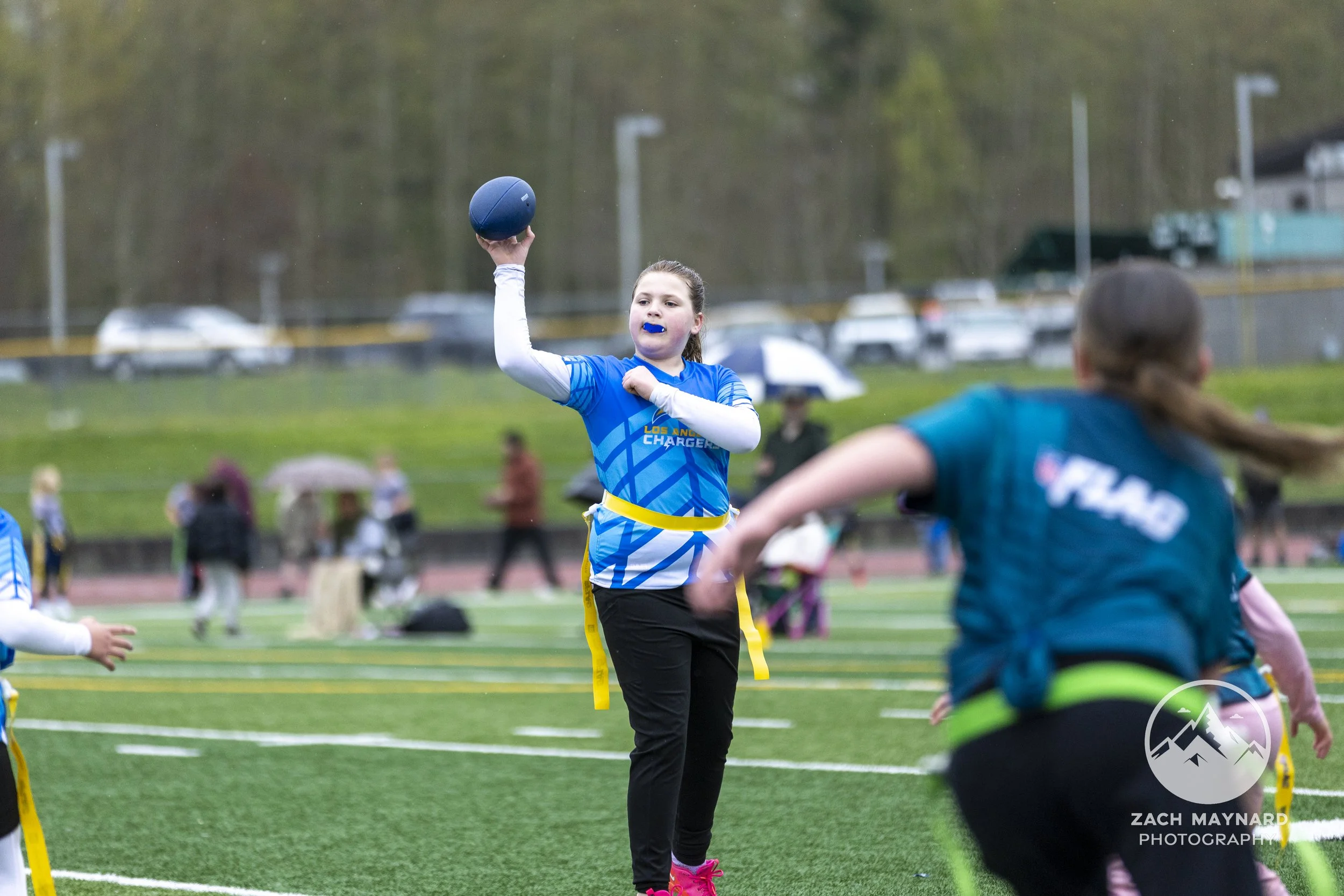 A young girl with a blue Los Angeles Chargers sports jersey and black pants, holding a blue ball in her right hand, participating in a track and field event. She has a blue mouthguard, her hair is tied back, and she is on a sports field with other children, adults, and trees in the background.