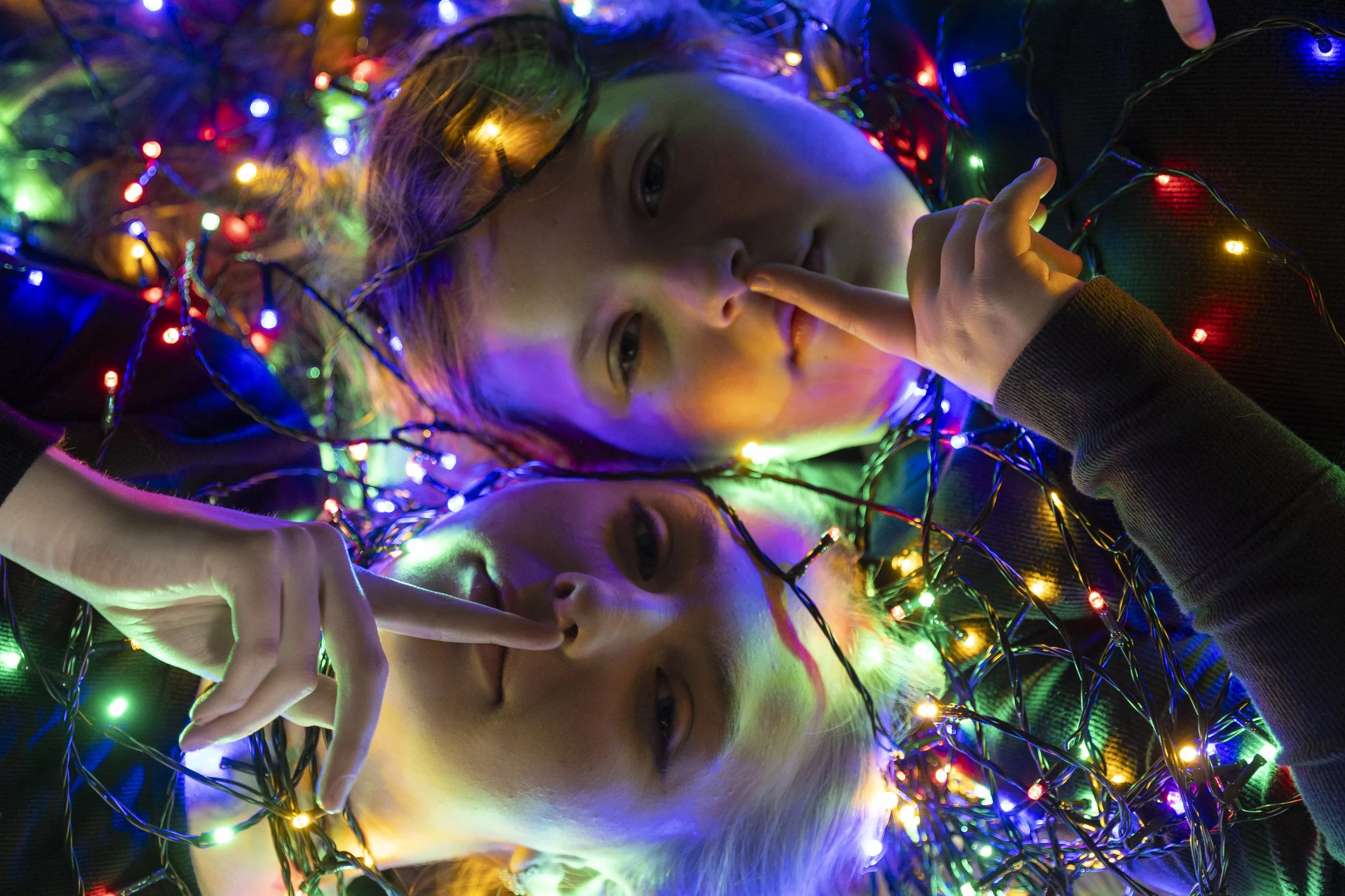 Two people lying down surrounded by colorful Christmas lights, with one person touching their lips with a finger and the other person holding a finger near their nose.