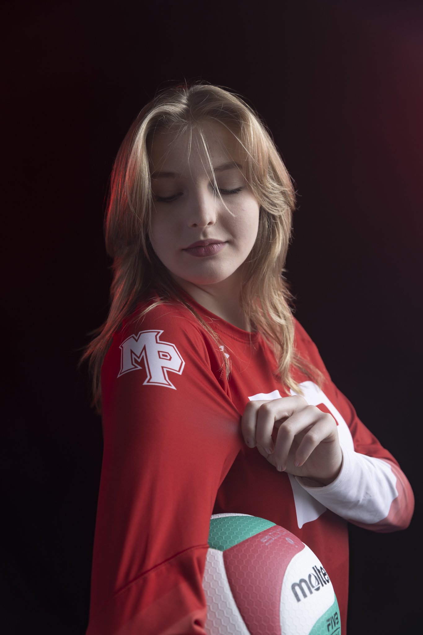 A young woman with long, wavy blonde hair and closed eyes, wearing a red sports jersey with white and light blue accents and holding a volleyball, standing against a dark background.