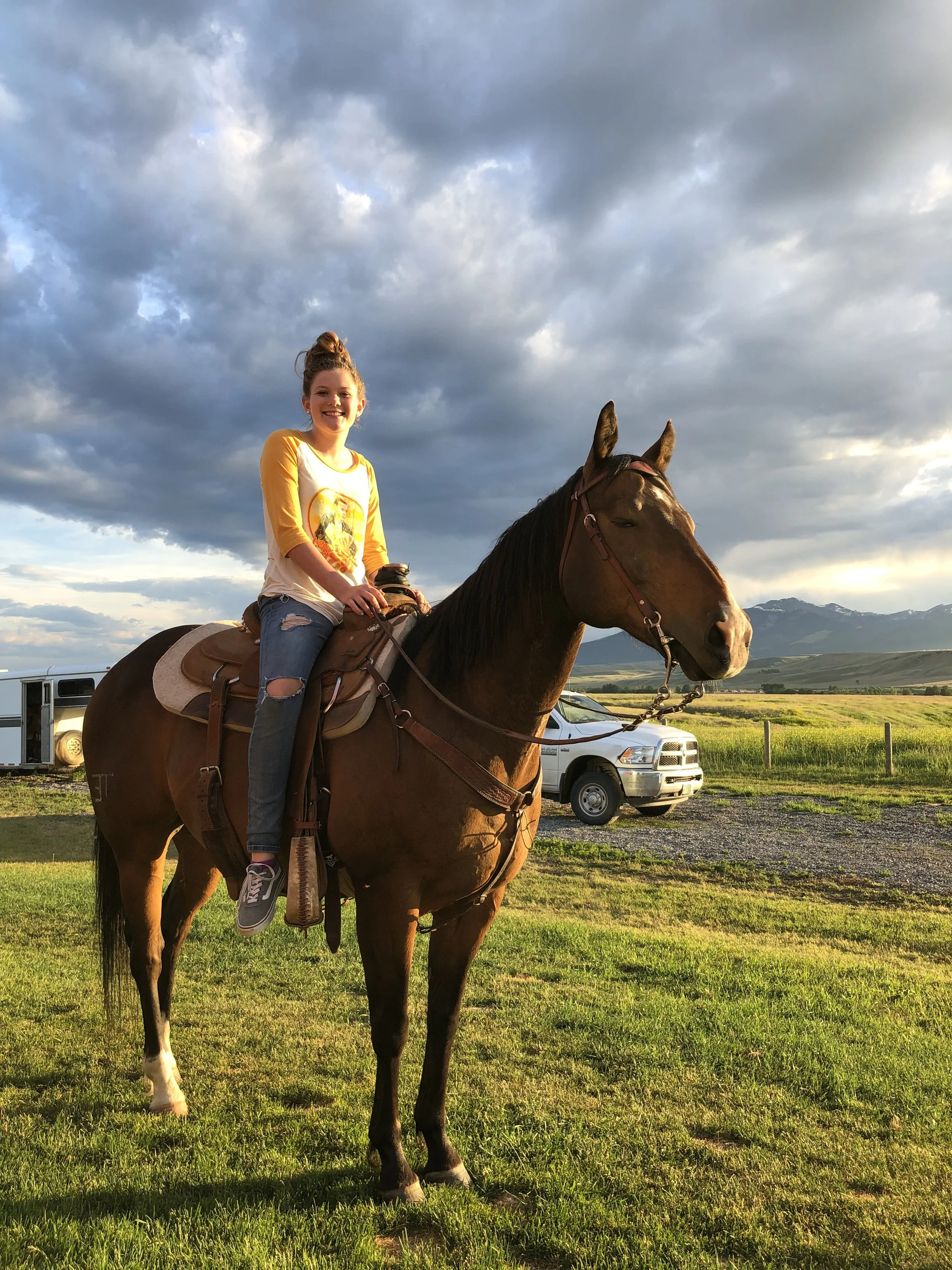 a girl in a yellow shirt and jeans sitting on a horse in front of a white car with grass and clouds in the background