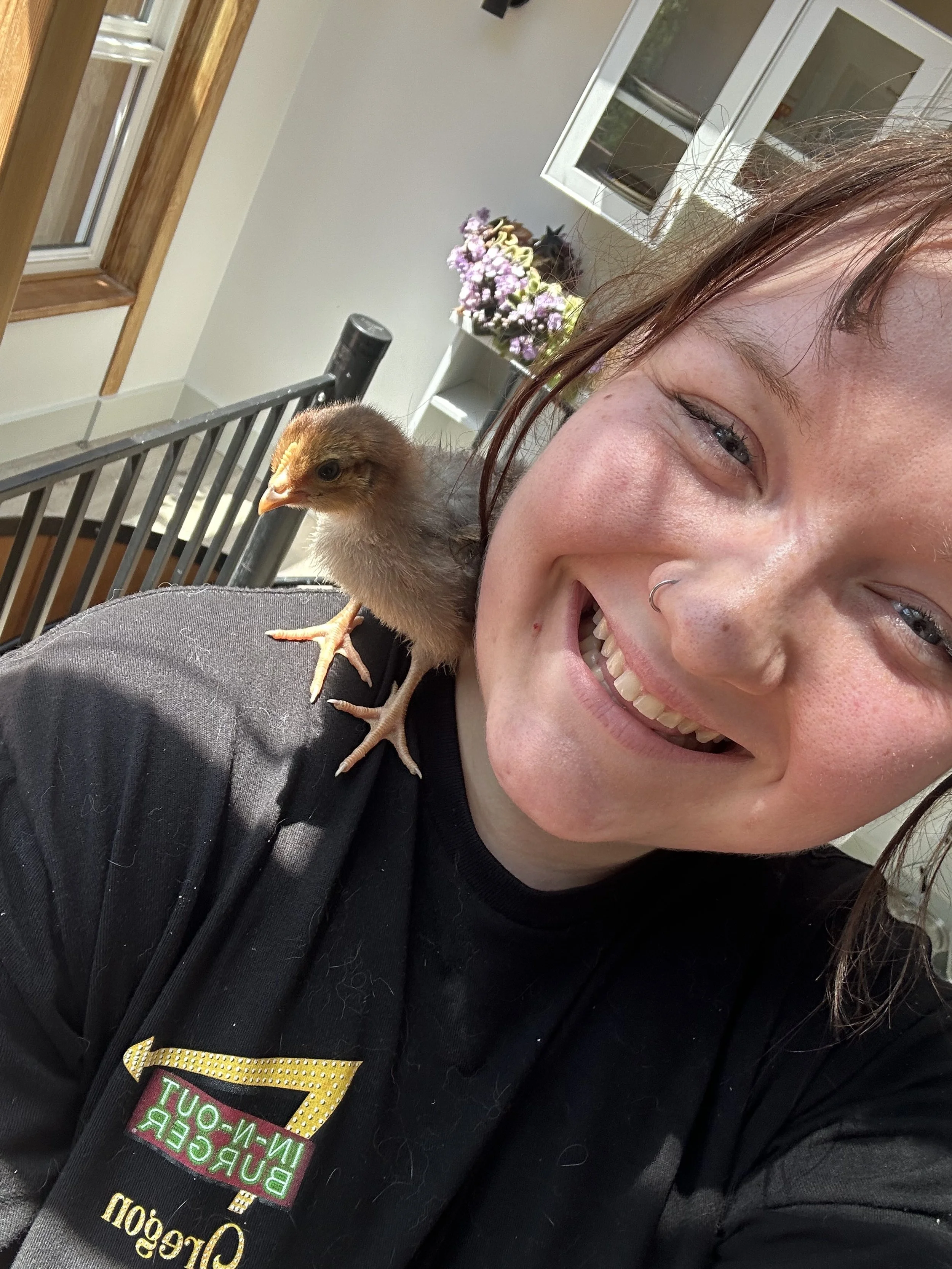 a woman in a black shirt smiling at the camera with a baby chicken sitting on her shoulder