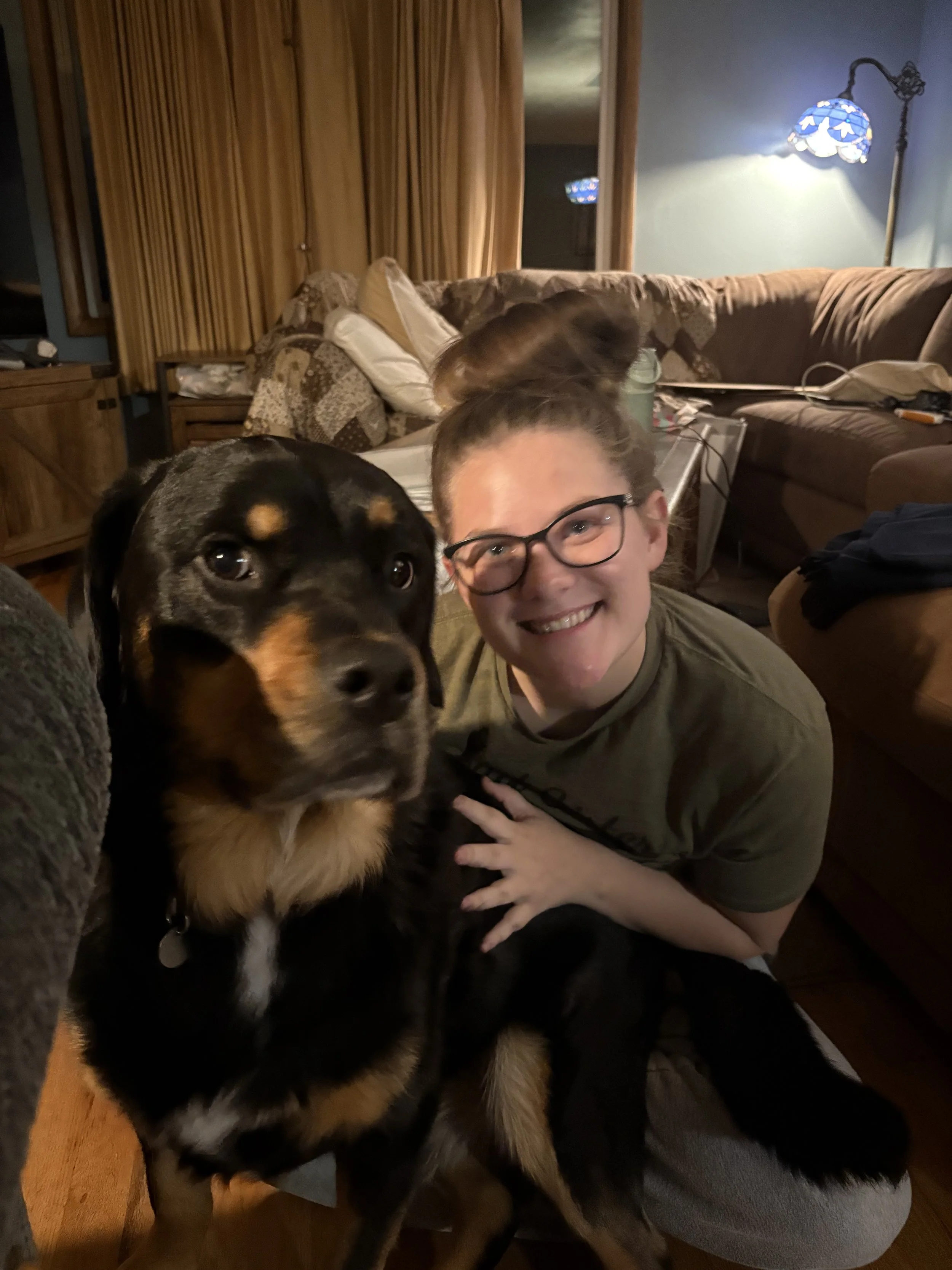 a girl in a gray shirt and glasses smiling and posing next to a rottweiler