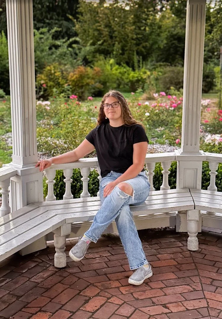 A young woman with glasses and curly hair sitting on a white wooden gazebo bench in a garden with blooming flowers and green trees in the background.