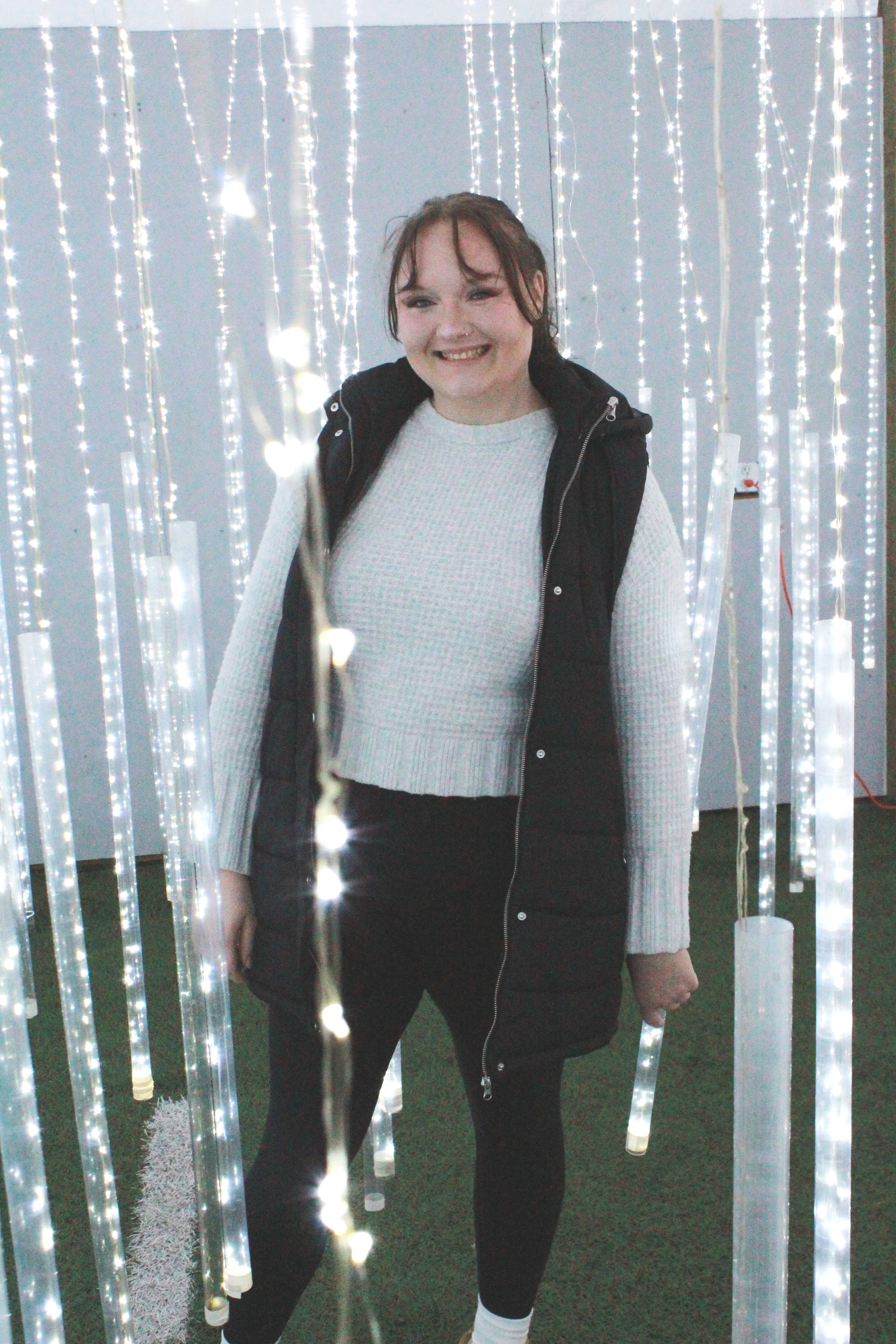 A young woman smiling and standing among hanging LED light strings in an indoor setting with a white wall background.