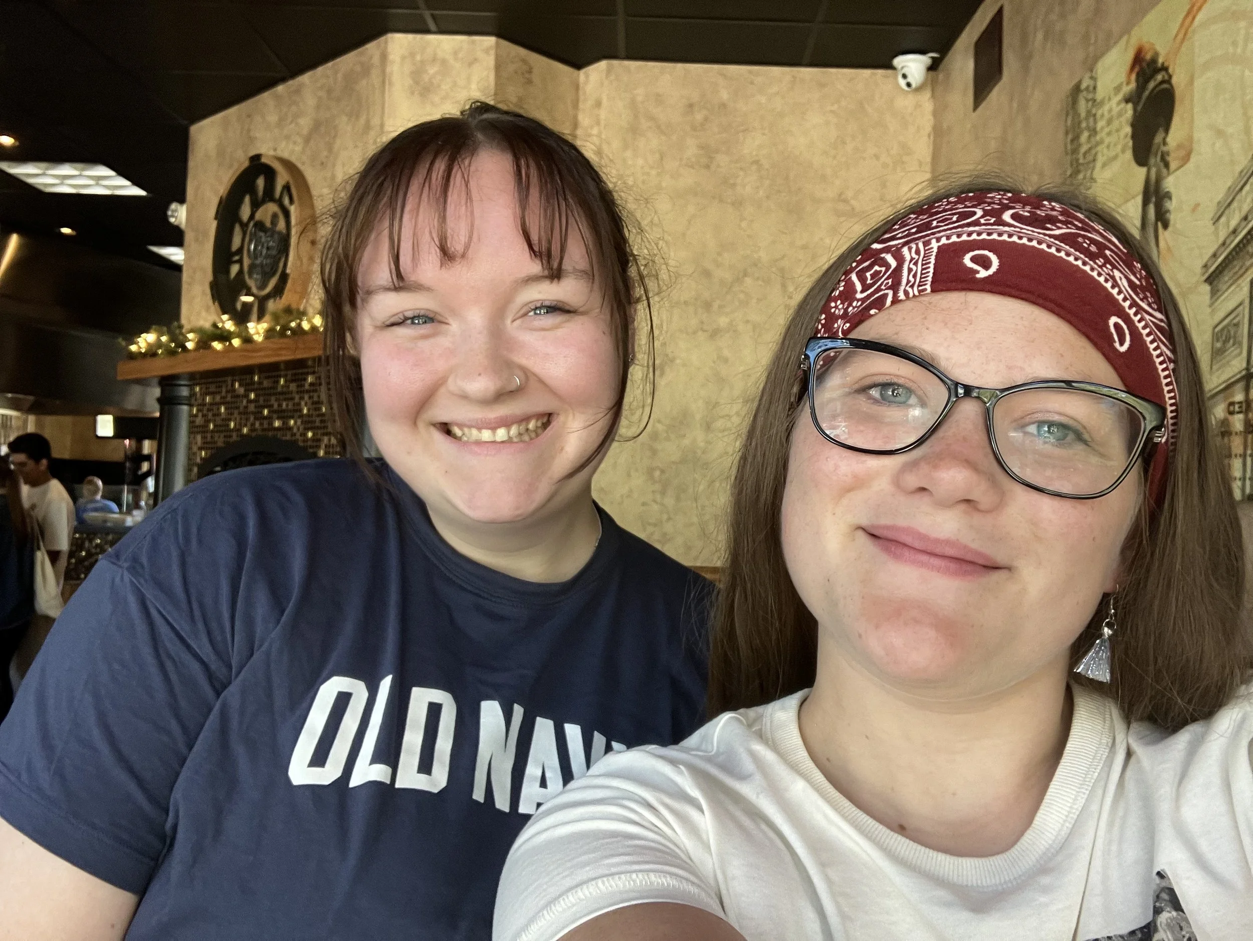 Two women smiling, taking a selfie inside a restaurant or cafe with a decorative wall, clock, and some patrons in the background.