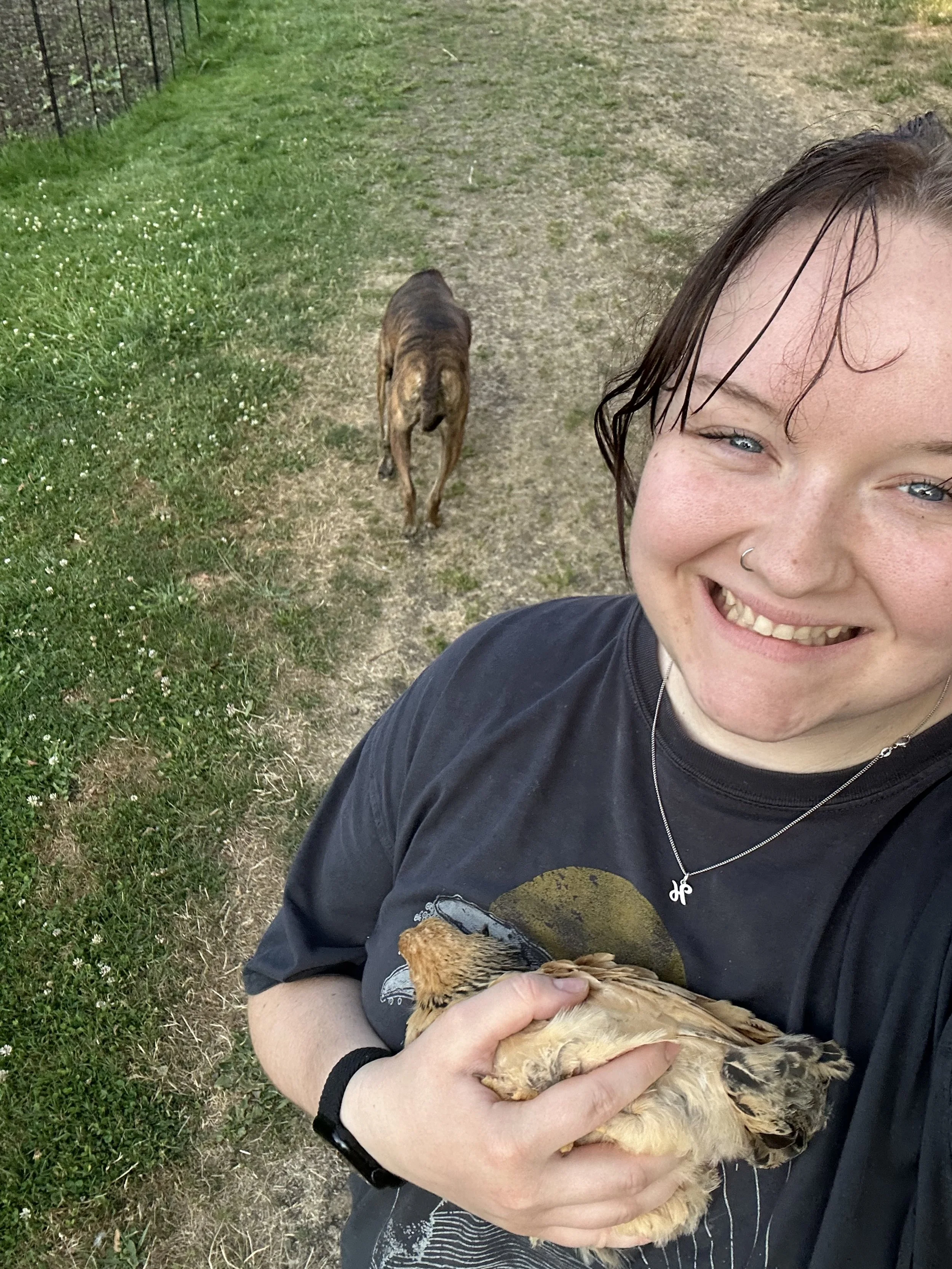 A girl with brown hair and a gray shirt standing and smiling at the camera while holding a chicken with a dog following behind her
