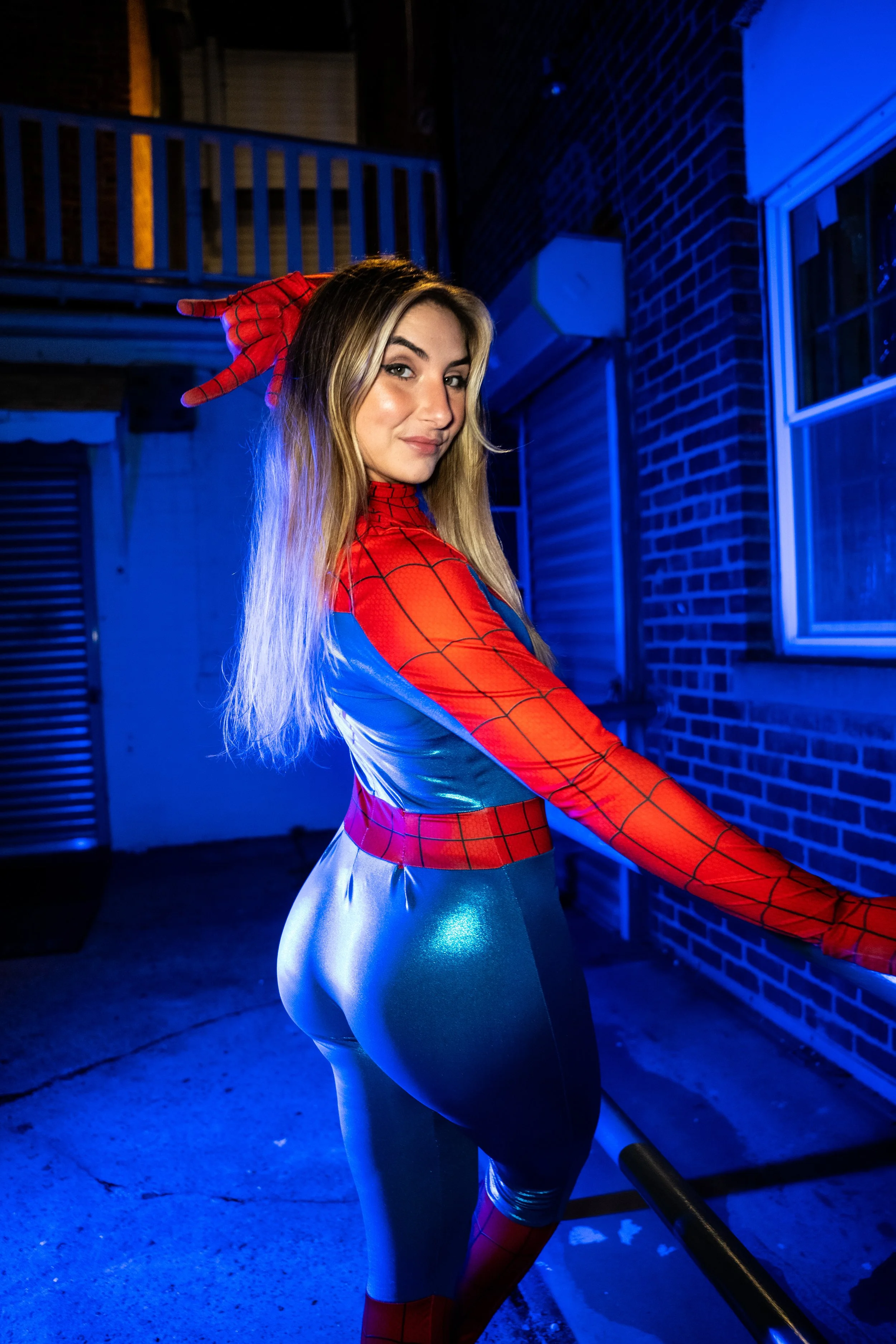 Lindsay poses in a red and blue Spider-Girl costume during a Halloween cosplay photoshoot in Patchogue, Long Island, illuminated by dramatic blue lighting against a brick building.