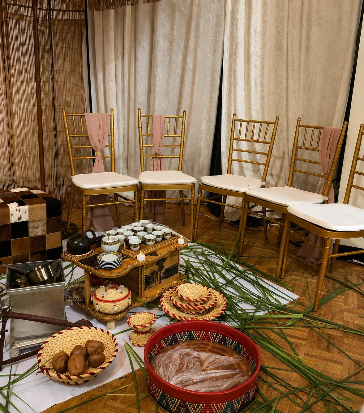 Chairs decorated with pink sashes, tableware including cups and plates, a variety of woven baskets, and green palm leaves arranged on the floor and table, in front of cream curtains.