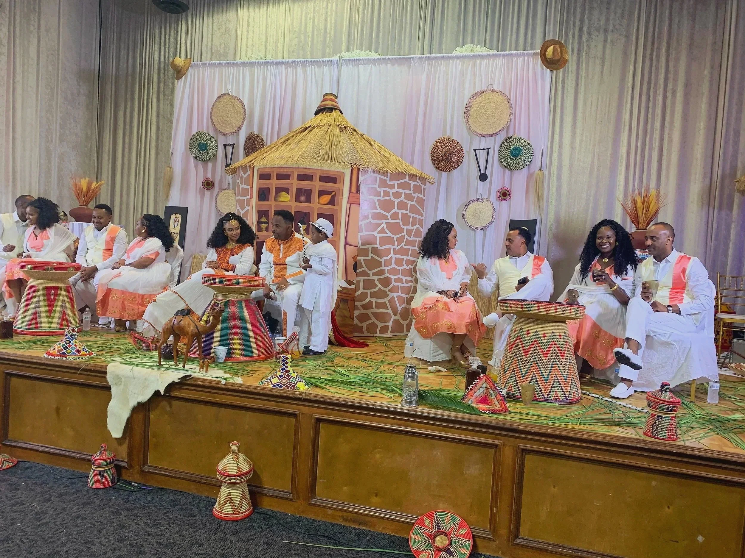 A group of people dressed in traditional African attire sitting on stage with a backdrop of white curtains decorated with woven baskets and hats, and Kenyan cultural artifacts and decorations in front.