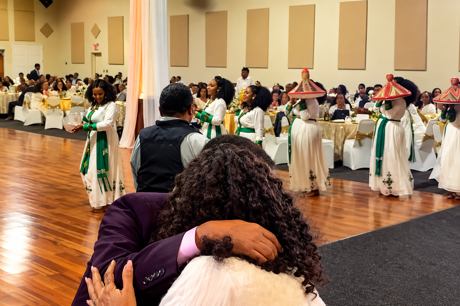 A woman and a man embrace in the foreground while a group of women in traditional dresses perform a dance in the background at a formal event with a large seated audience.