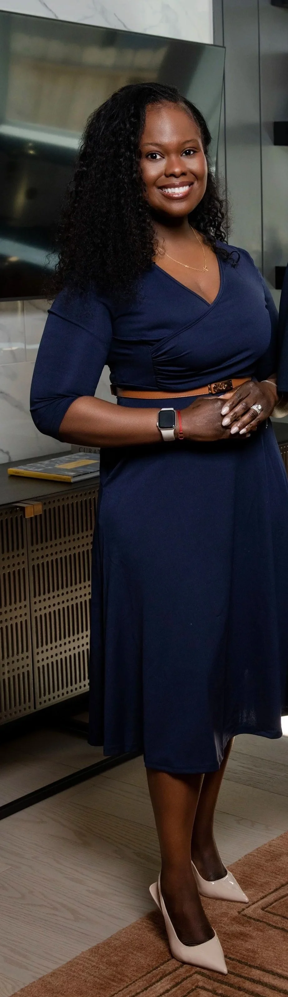 A smiling woman with dark curly hair is dressed in a navy blue dress, wearing beige high heels and a smartwatch, standing indoors with a wall-mounted TV and a wooden cabinet behind her.