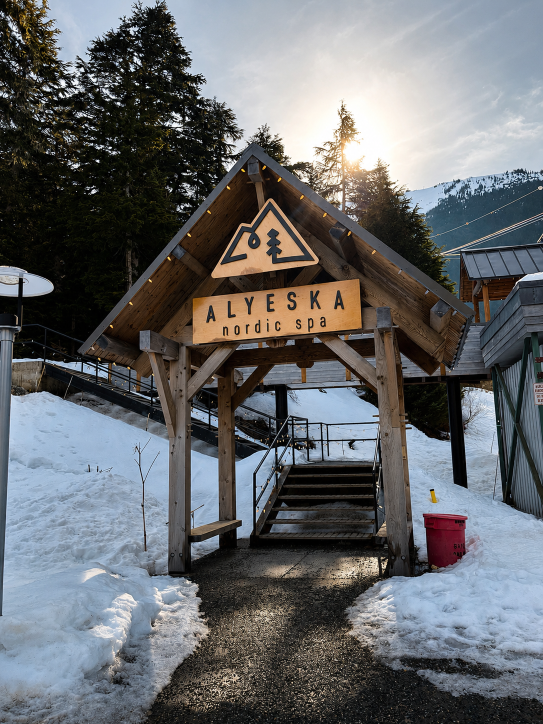 Wooden entrance sign for Alyeska Nordic Spa with snow-covered surroundings, pine trees, and mountain backdrop.