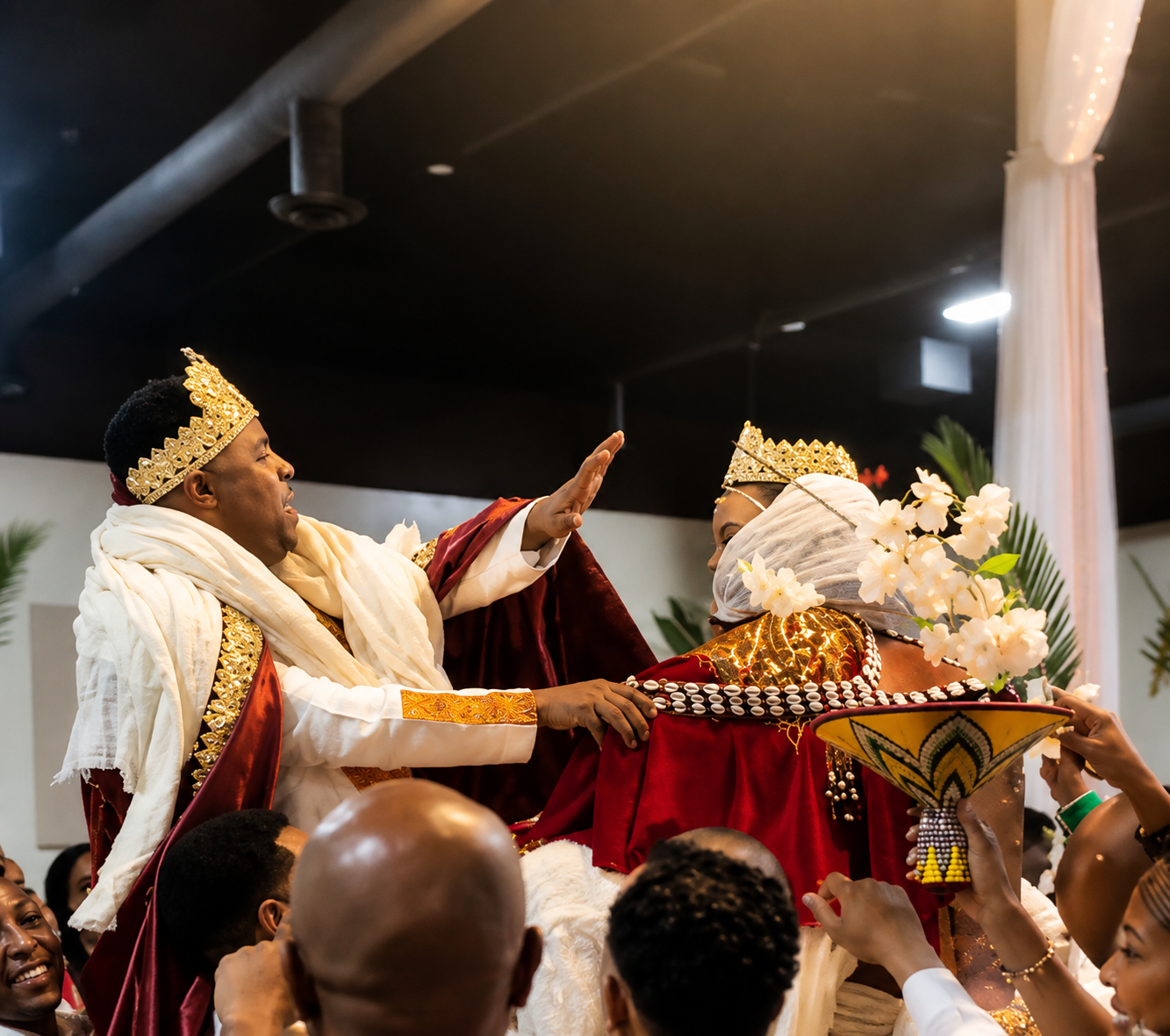 A traditional celebration with two people dressed in regal attire, wearing crowns, with one person raising their hand in a gesture. The scene includes floral and decorative elements, indicating a formal cultural or ceremonial event.