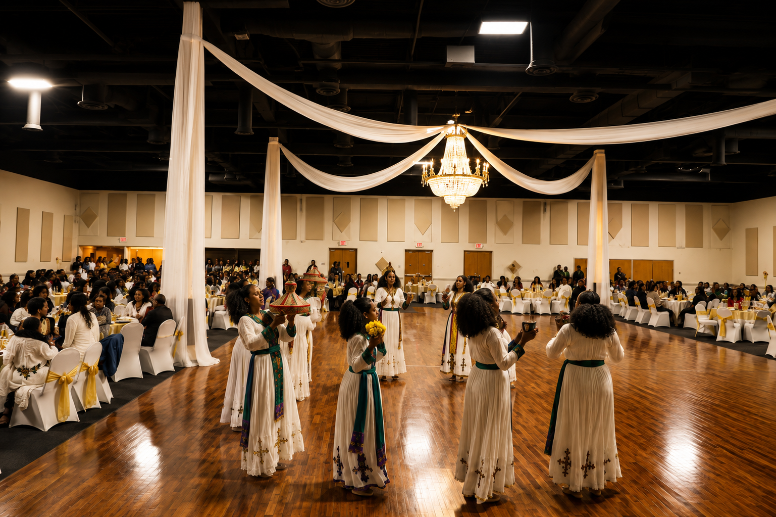 A group of women in traditional white dresses with green sashes performing a dance on a polished wooden floor during a cultural celebration in a banquet hall, decorated with white drapes and a large chandelier.
