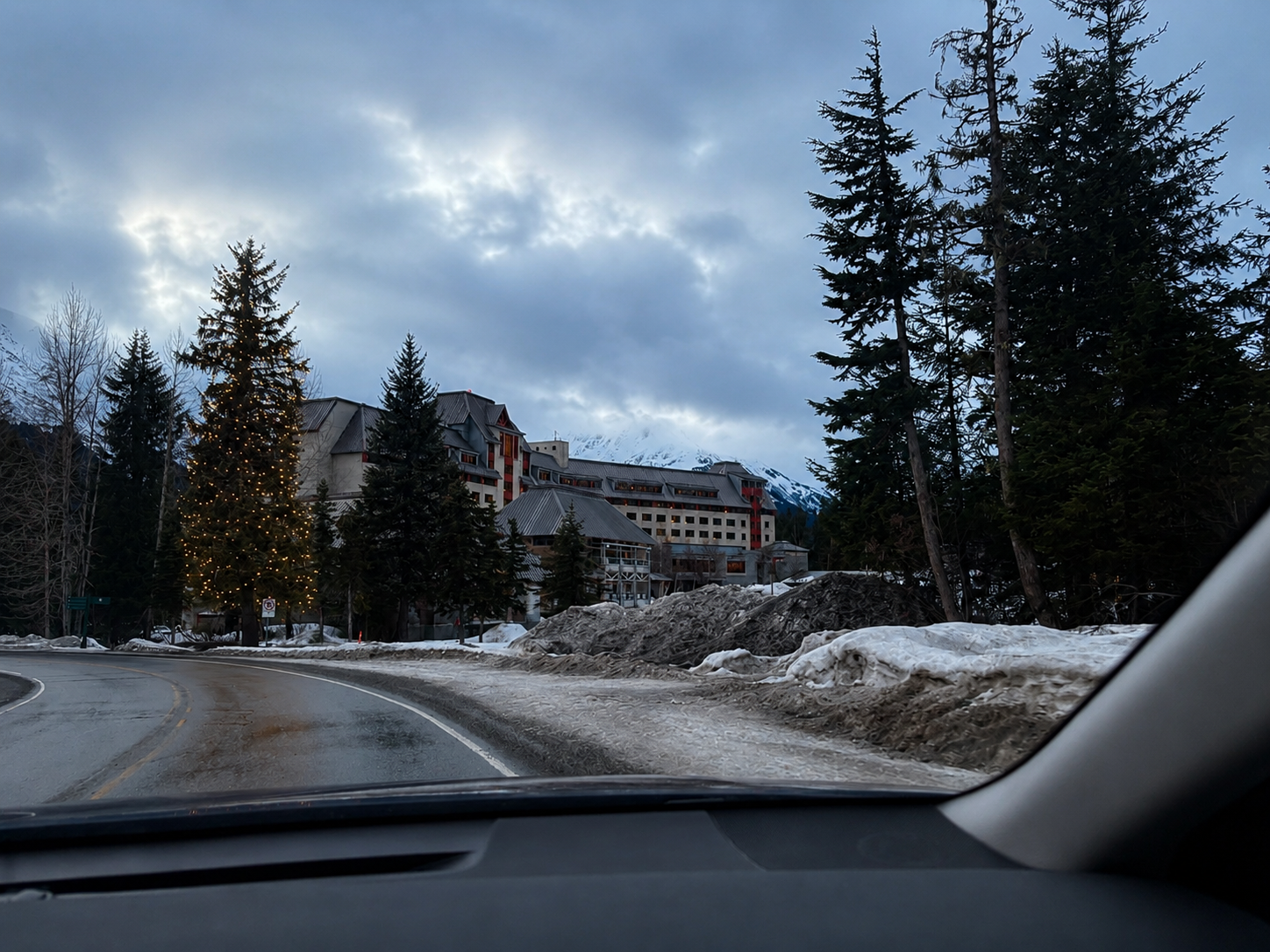 A snowy mountain resort building with a decorated Christmas tree and pine trees surrounding it, viewed from inside a car on a winding road with snow and ice.