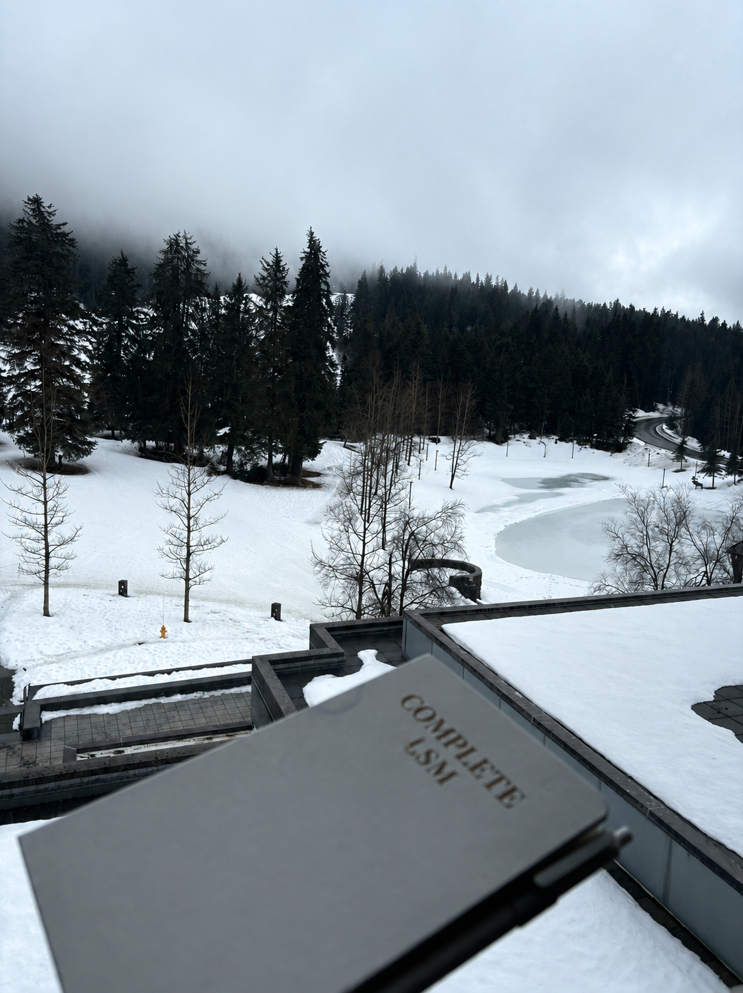 Snow-covered landscape with trees, a frozen pond, and a winding road in the distance, as seen from a building ledge with a 'Completer U.S.M' notepad in the foreground.