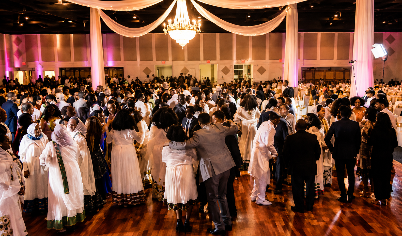 People gathered on a dance floor at an indoor event, with a large chandelier, pink lighting, and decorated drapery ceiling.