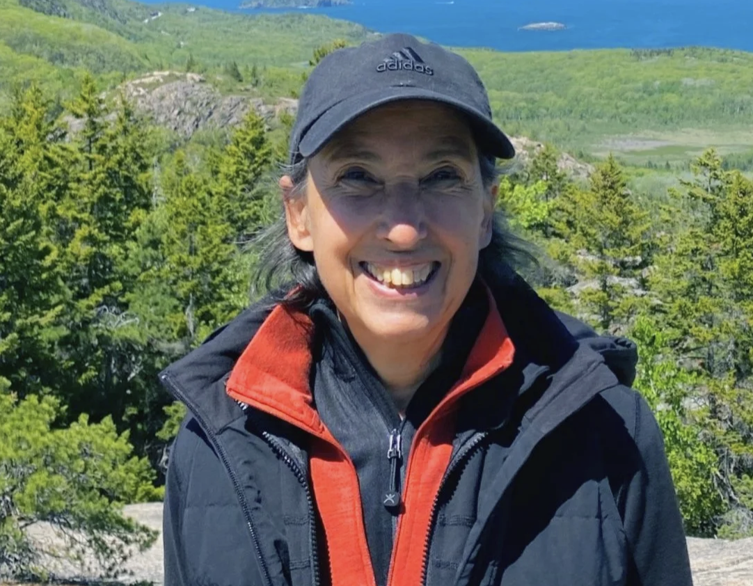 A smiling woman wearing a black cap, black and red outdoor jacket, standing in a green mountainous landscape with trees and a blue sky.