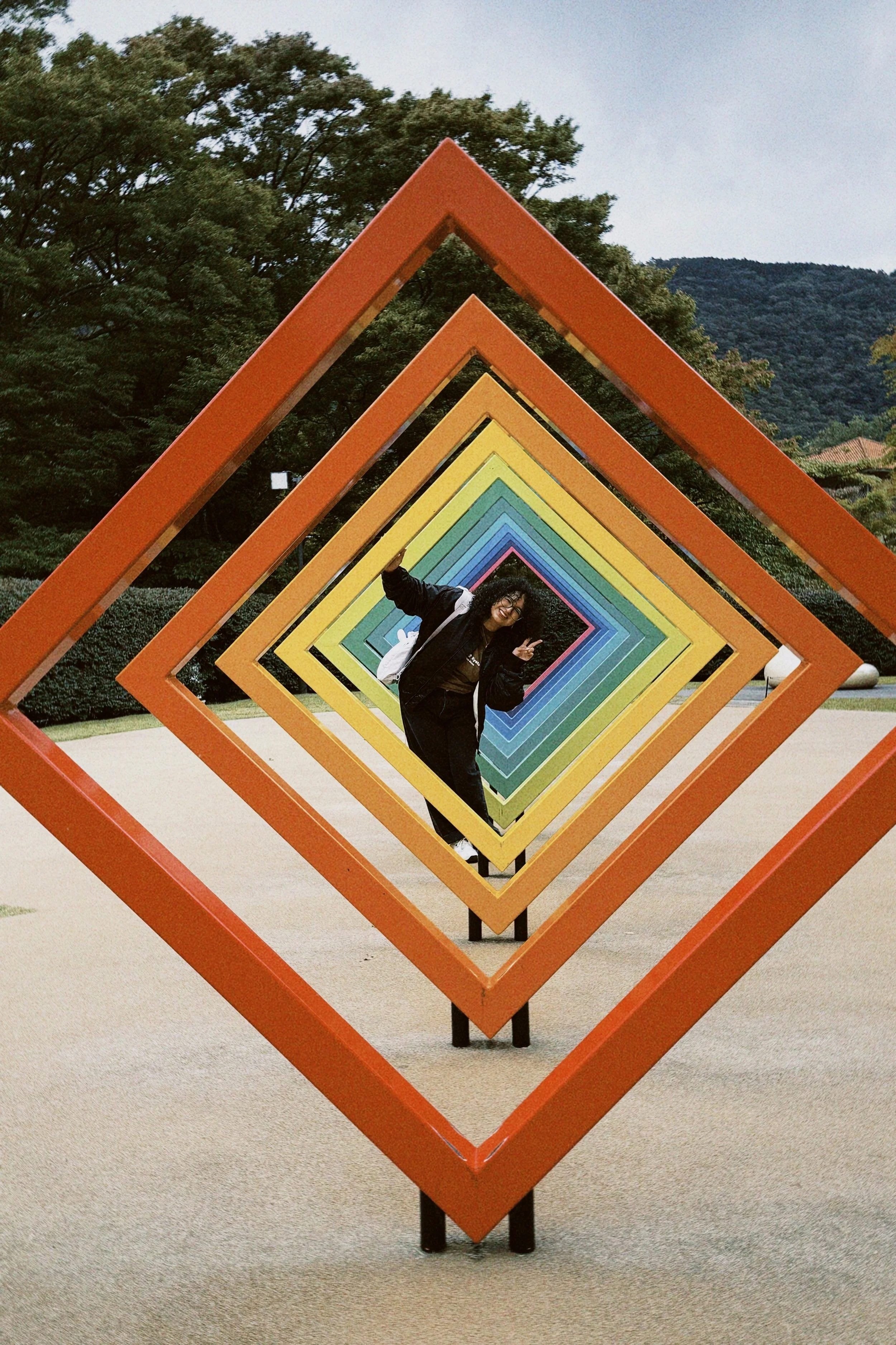 A person standing smiling inside a series of colorful square frames arranged in a tunnel-like installation outdoors, with trees and mountains in the background.