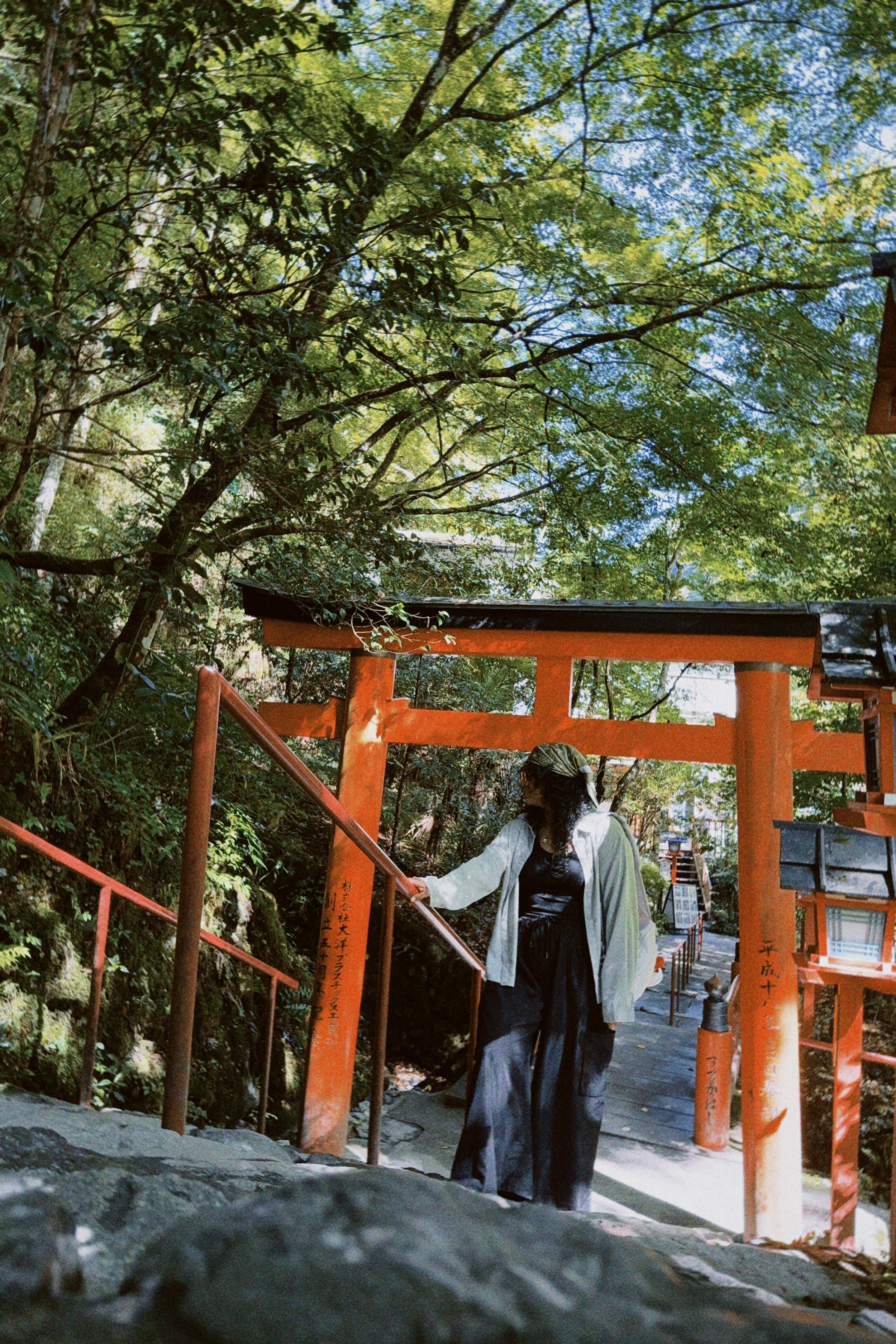 A woman wearing black clothing, a gray jacket, and a silk headscarf, standing on a stone step, holding a red railing at a traditional Japanese shrine with orange gates surrounded by lush greenery and trees.