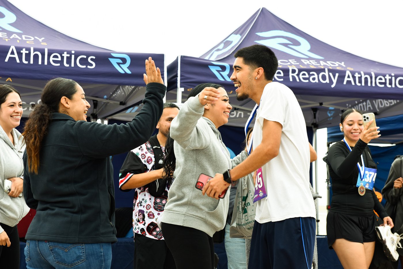 A group of runners celebrating at a finish line, smiling and high-fiving, with tents in the background that read 'Race Ready Athletics'.