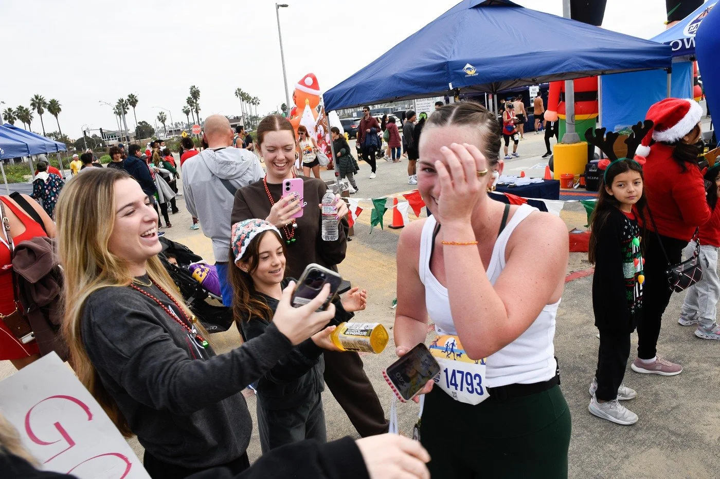 A group of women and girls at a holiday-themed outdoor event, with some wearing Christmas accessories like Santa hats and reindeer antlers, smiling and taking photos.