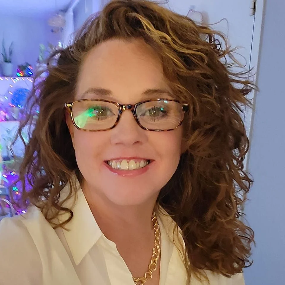 A smiling woman with curly red hair wearing glasses, a white shirt, and a gold necklace, standing indoors near a decorated Christmas tree.