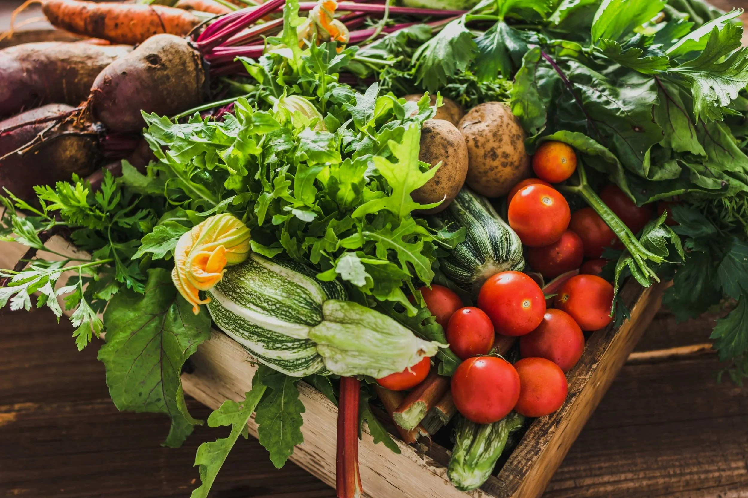 Fresh vegetables including cherry tomatoes, zucchinis, potatoes, carrots, and leafy greens in a wooden crate.