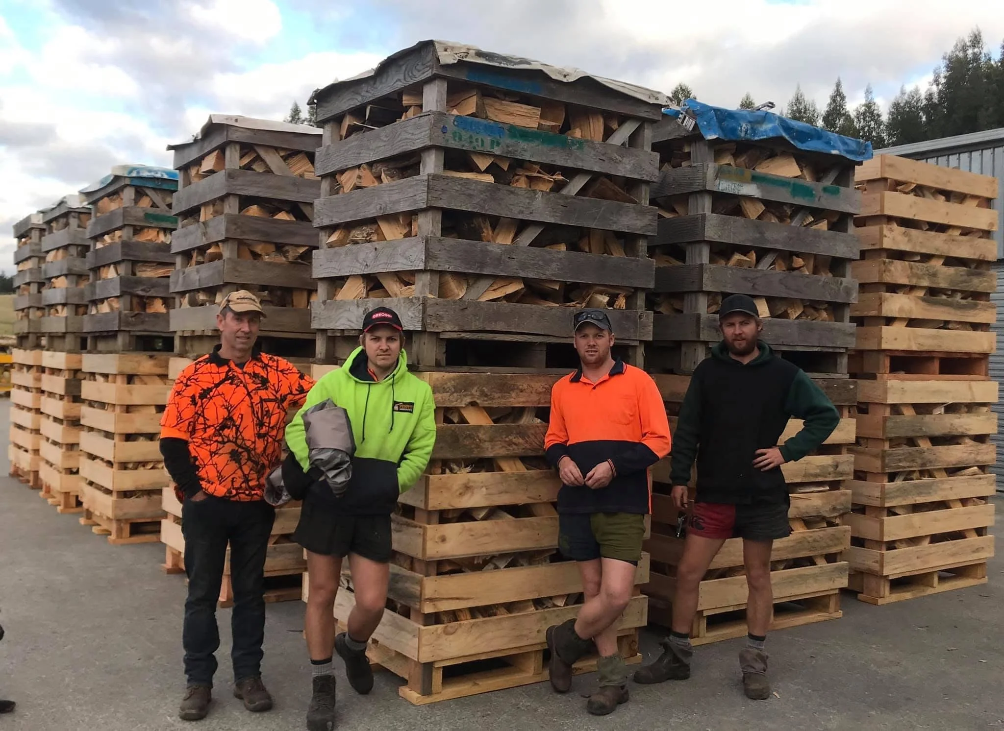 Four men standing in front of a large stack of cut firewood on pallets outdoors.