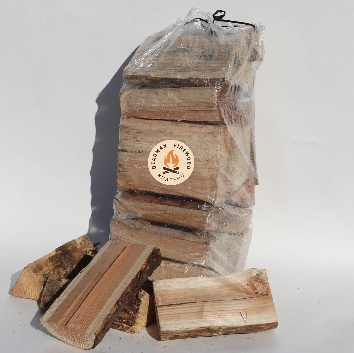 A bundle of firewood wrapped in plastic with a Deadman Firewood logo sticker, placed on a white surface with some loose wood pieces surrounding it.