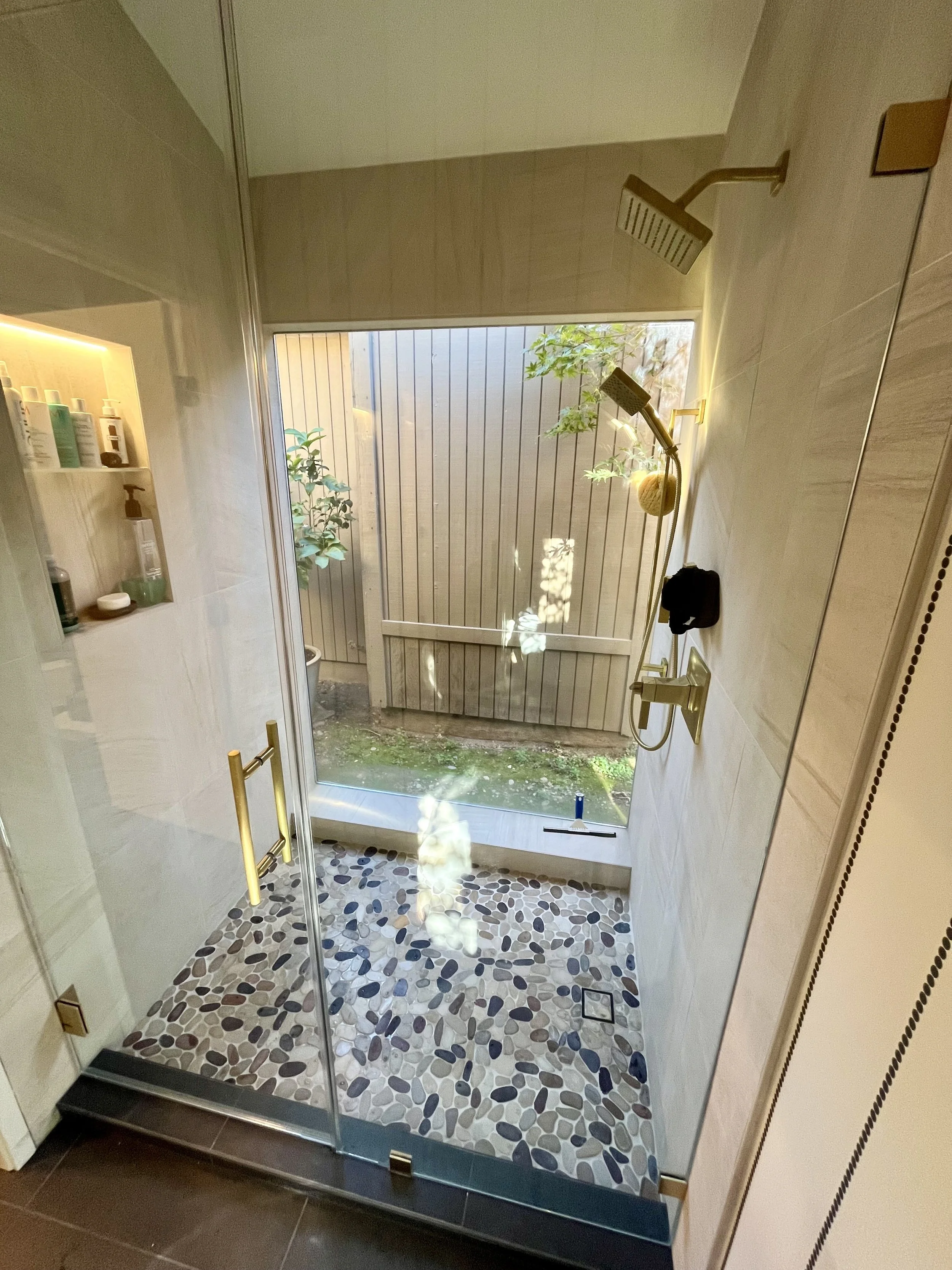 Shower area with glass door, pebble tile floor, dual showerheads, a bookshelf with toiletries, and outdoor plants visible through the window.