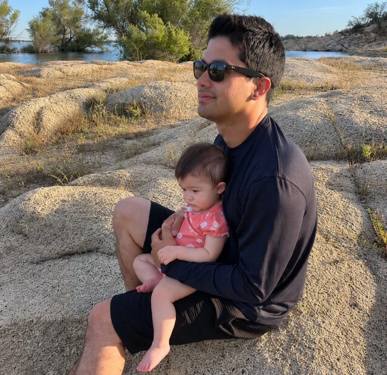 A young man wearing sunglasses and a navy long sleeve shirt sitting on rocks by a body of water, holding a little girl in a pink floral dress.