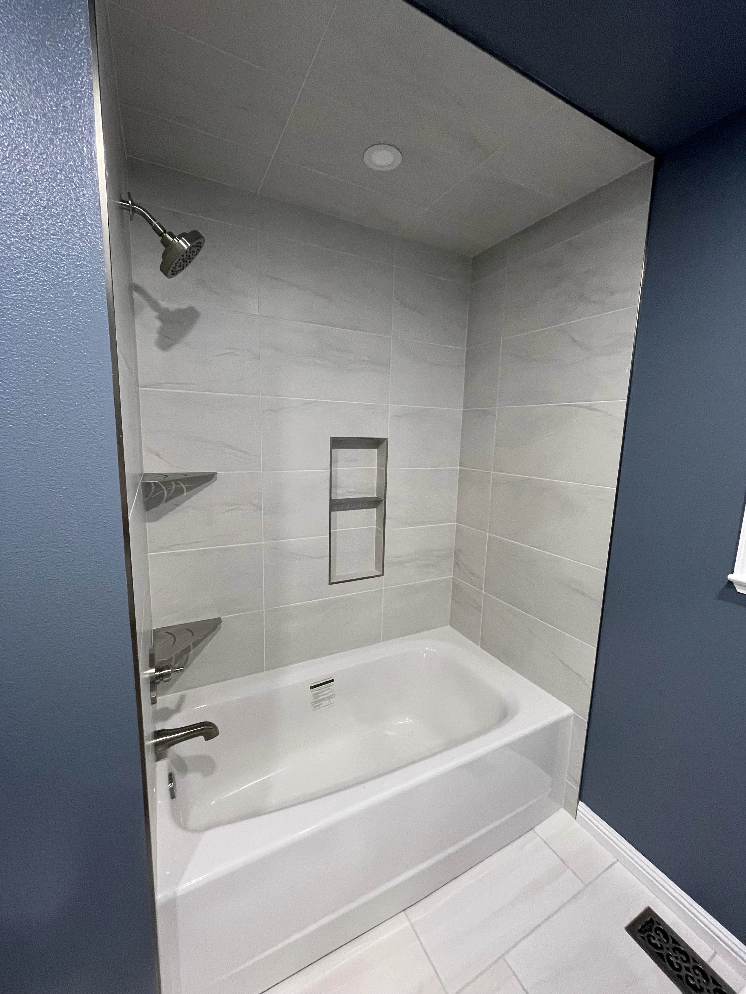 A corner shower with a white bathtub, a silver faucet, tiled walls, a built-in shelf, and a vent on the ceiling.