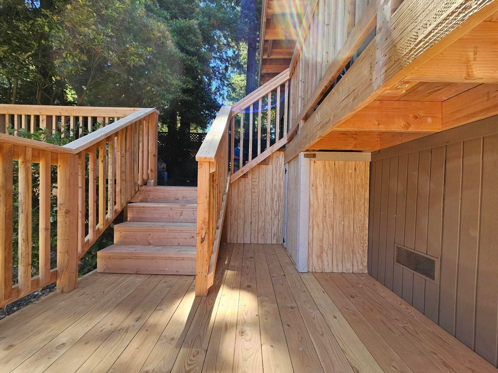 Wooden deck with stairs and railings, outdoor environment with trees in the background, sunlight creating lens flare.