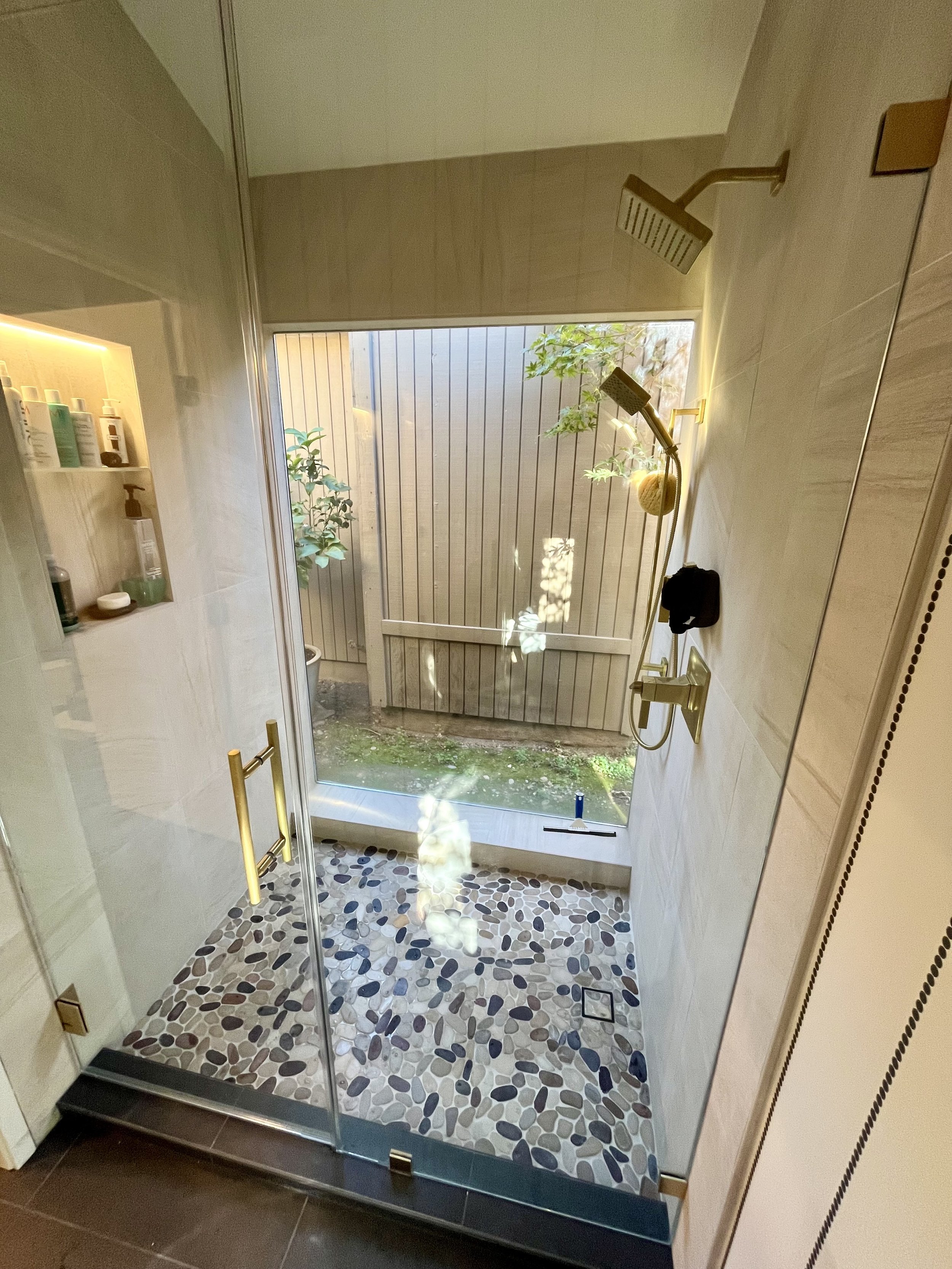 Bathroom shower with glass door, pebble-patterned floor, and a small outdoor garden visible through a window at the back of the shower.