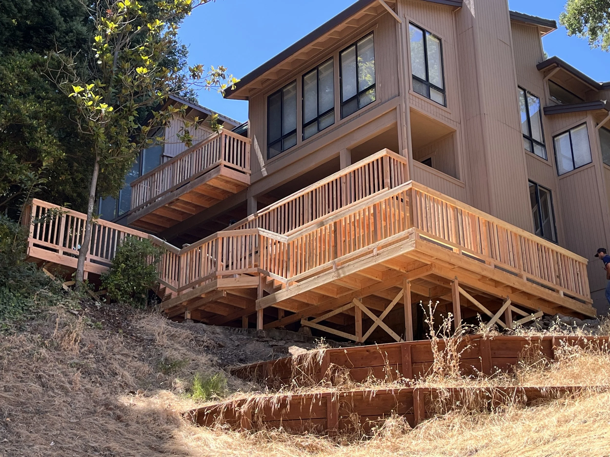 A multi-level house with a wooden exterior and extensive glass windows is built on a hillside among trees. It features decks with wooden railings on different floors and has a sloped roof. The sky is clear and blue.