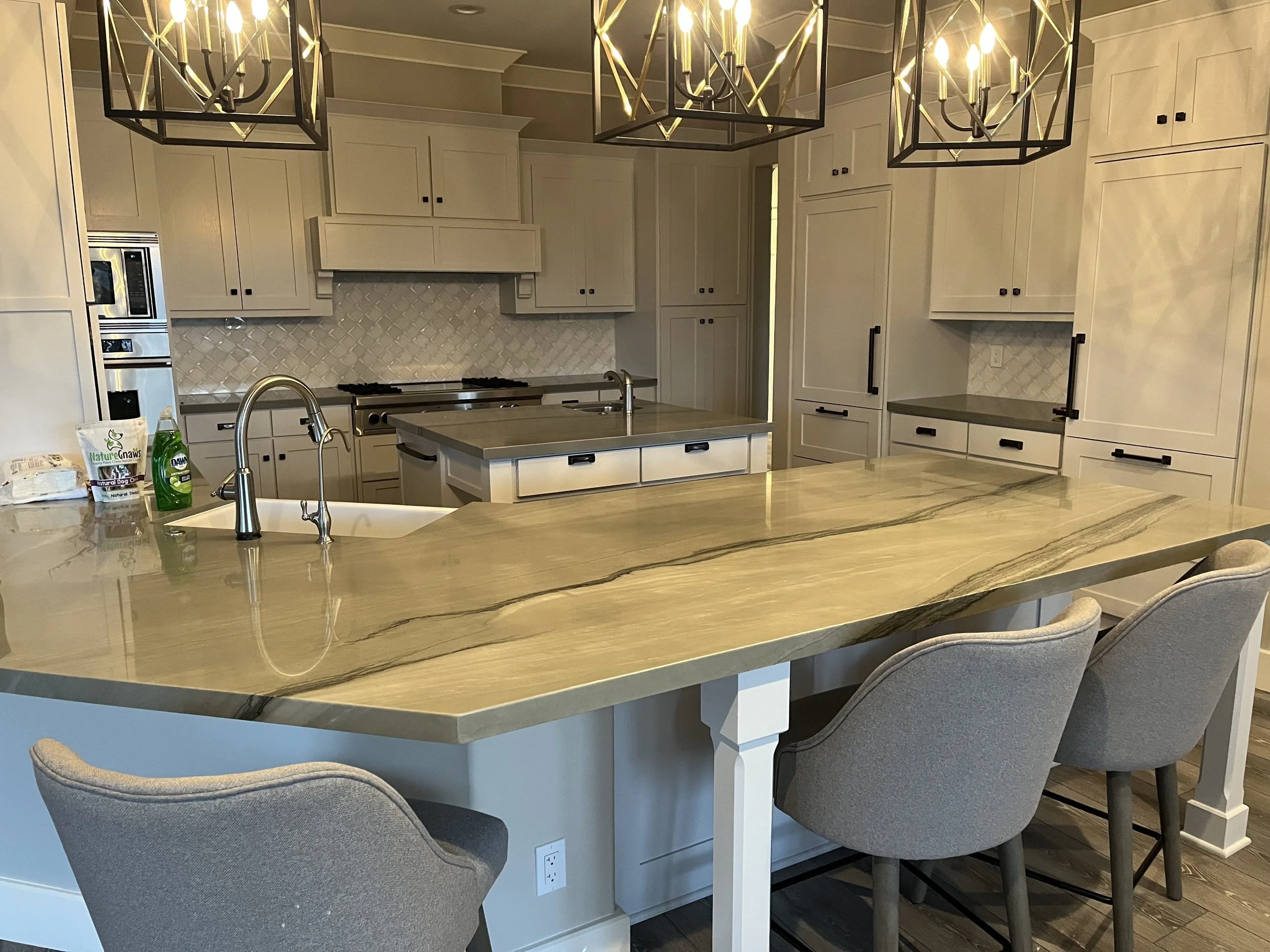 Modern kitchen with white cabinets, a marble island, and black hardware. Overhead, three geometric black and gold light fixtures hang. The setting includes a sink, a stove, and some bottles on the counter.
