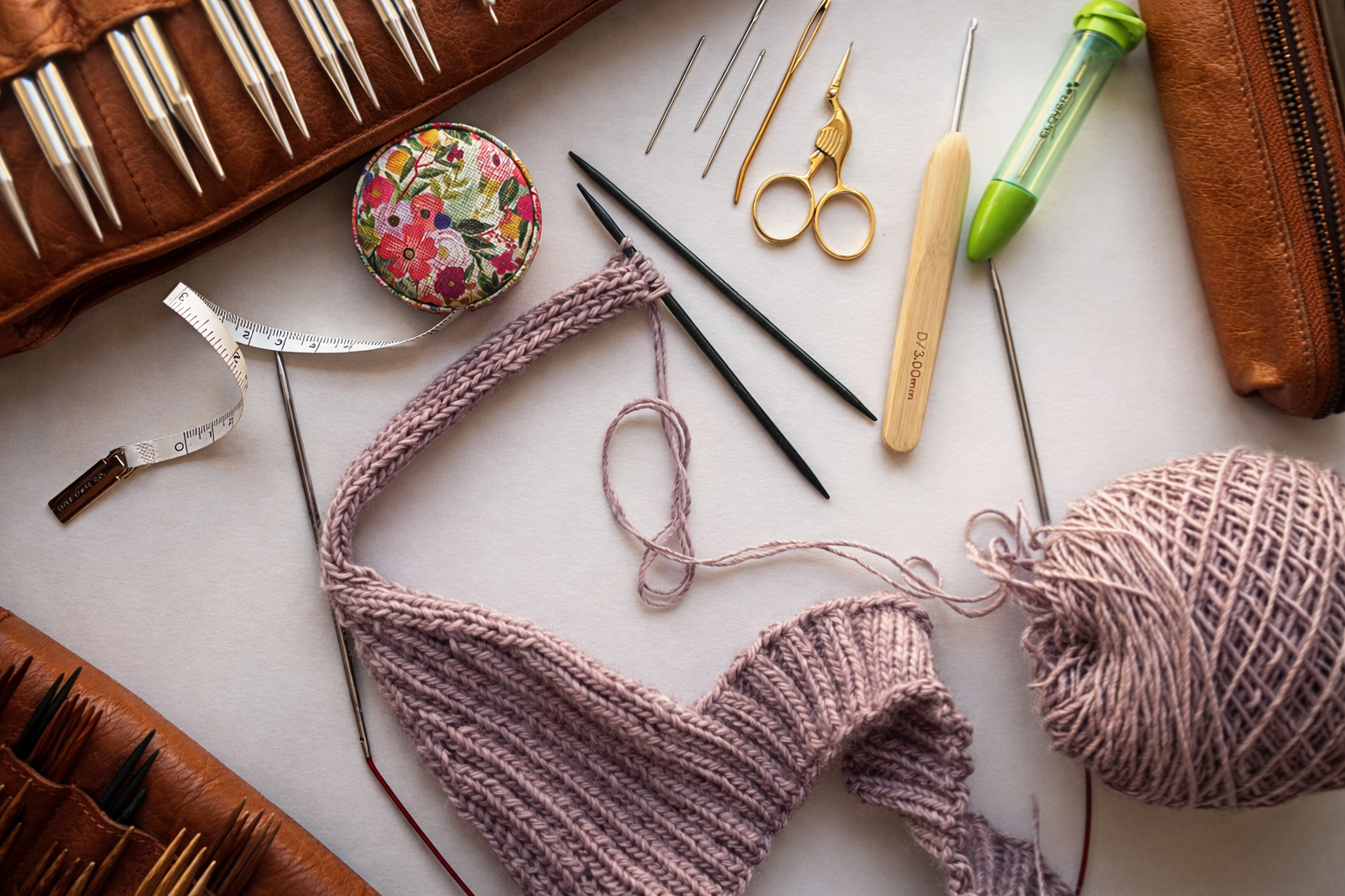 Knitting yarn, knitting needles, and sewing tools arranged on a white surface, with a sewing kit and crochet project in progress.