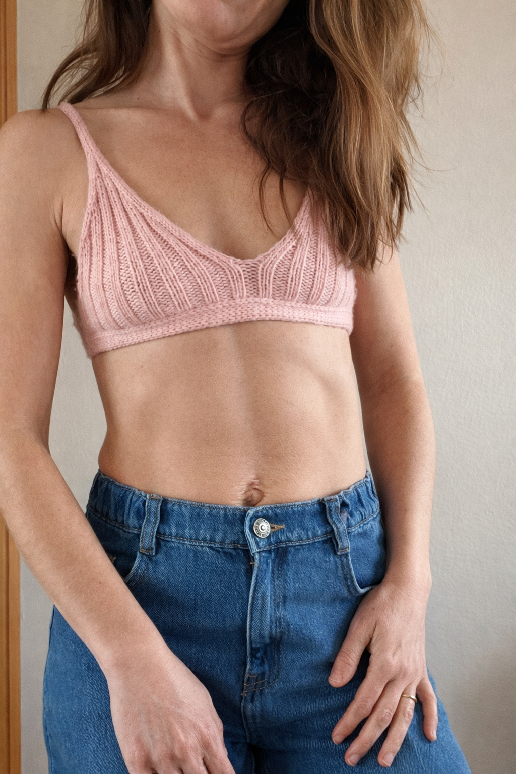 A woman wearing a pink knitted crop top and blue jeans, standing indoors against a plain wall.