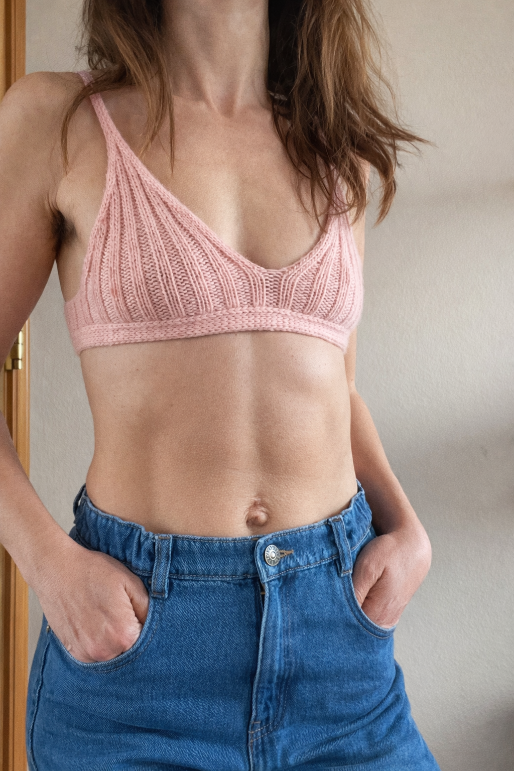 A woman wearing a pink knitted bralette and blue high-waisted jeans with hands in pockets, standing indoors against a neutral background.