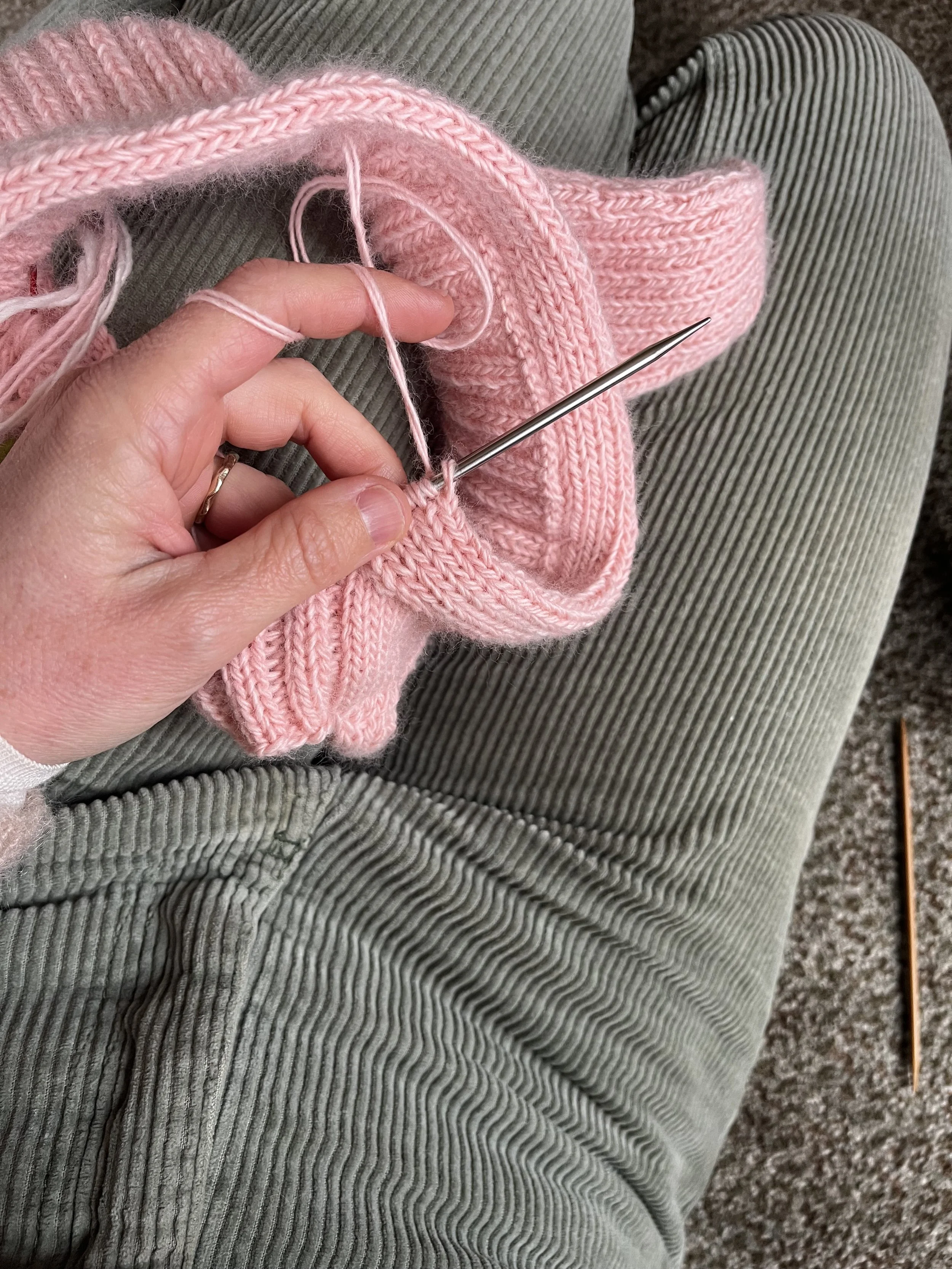A person knitting a pink yarn item using a metal knitting needle, sitting on a green corduroy fabric surface.